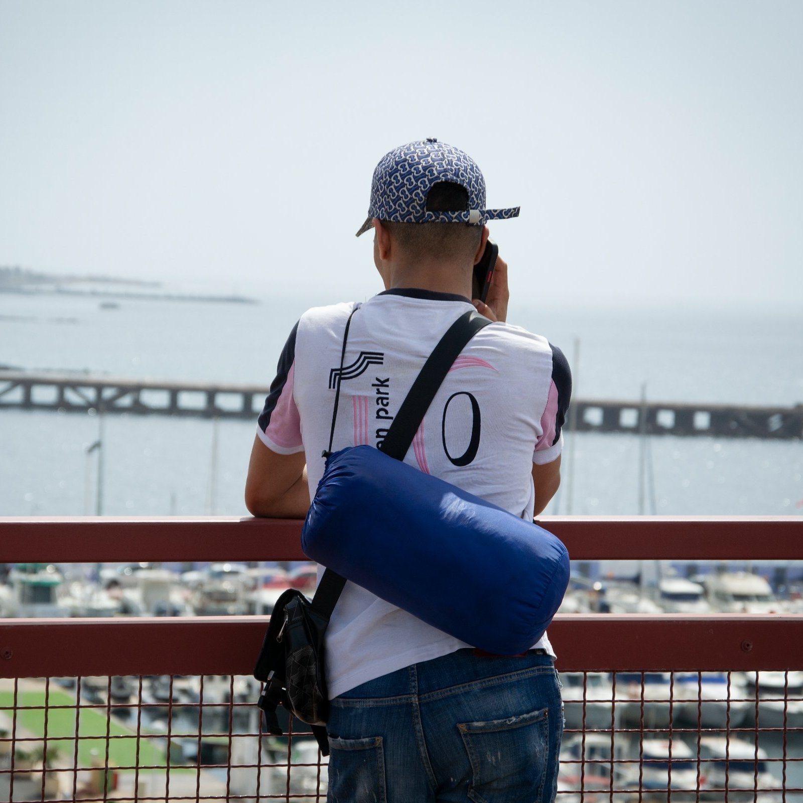 Dans cette image, un homme se tient de dos, regardant au loin vers la mer. Il est vêtu d'un t-shirt blanc avec des accents colorés et porte une casquette à motifs. Sur son épaule, il a un petit sac bleu. L'arrière-plan montre un port avec des bateaux amarrés et une jetée qui s'étend dans l'eau, créant une atmosphère tranquille. Le ciel est clair, suggérant une journée ensoleillée. L'homme semble concentré sur ce qu'il observe, profitant de la vue sur l'horizon maritime.