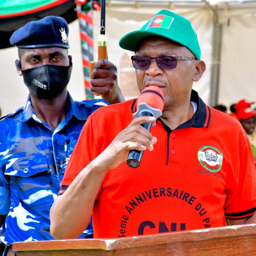 L'image montre un homme qui prend la parole devant un public. Il porte un t-shirt rouge avec le texte "ANNIVERSAIRE DU PARTI" et une casquette verte. Il tient un micro et semble s'adresser à l'assemblée avec passion. À l'arrière-plan, on peut apercevoir des gens assis, certains portant des vêtements colorés. Un agent de sécurité en uniforme bleu est visible à côté de lui, ajoutant un élément de protection ou d'autorité à l'événement. L'atmosphère semble festive et engagée.