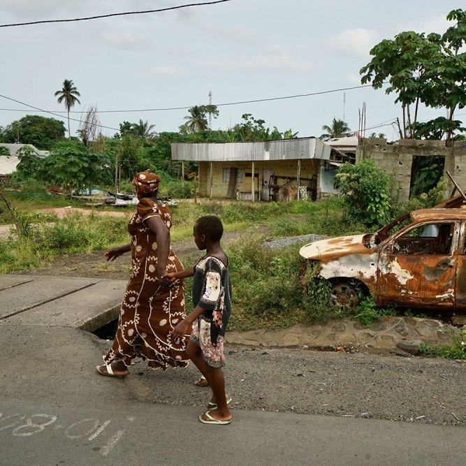 L'image montre une scène d'une rue dans un environnement rural ou semi-urbain. Sur le trottoir, une femme vêtue d'une longue robe ornée de motifs blancs marche aux côtés d'un jeune garçon. Ils avancent côte à côte, probablement en direction d'un endroit précis. En arrière-plan, on peut voir un paysage verdoyant avec des arbres, ainsi que des bâtiments partiellement construits, évoquant une ambiance de quartier en développement. À droite, un vieux véhicule, rouillé et en piteux état, est abandonné sur le côté de la route, entouré de végétation. Le ciel est nuageux, suggérant qu'il pourrait faire chaud et humide. L'ensemble dégage une atmosphère à la fois paisible et un peu nostalgique.