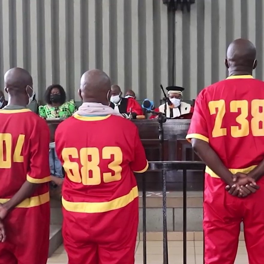 L'image montre un groupe de personnes dans un tribunal. Quatre hommes, portant des uniformes rouges numérotés au dos (337, 404, 683, 238, et 72), se tiennent debout, de dos, face à un juge et à un panel de personnes assises en face d'eux. L'atmosphère est sérieuse, et tous les participants portent des masques. Les murs du tribunal sont rayés, et on peut voir des membres de l'auditoire, y compris des femmes en vêtements colorés. L'ambiance générale évoque une situation judiciaire formelle.