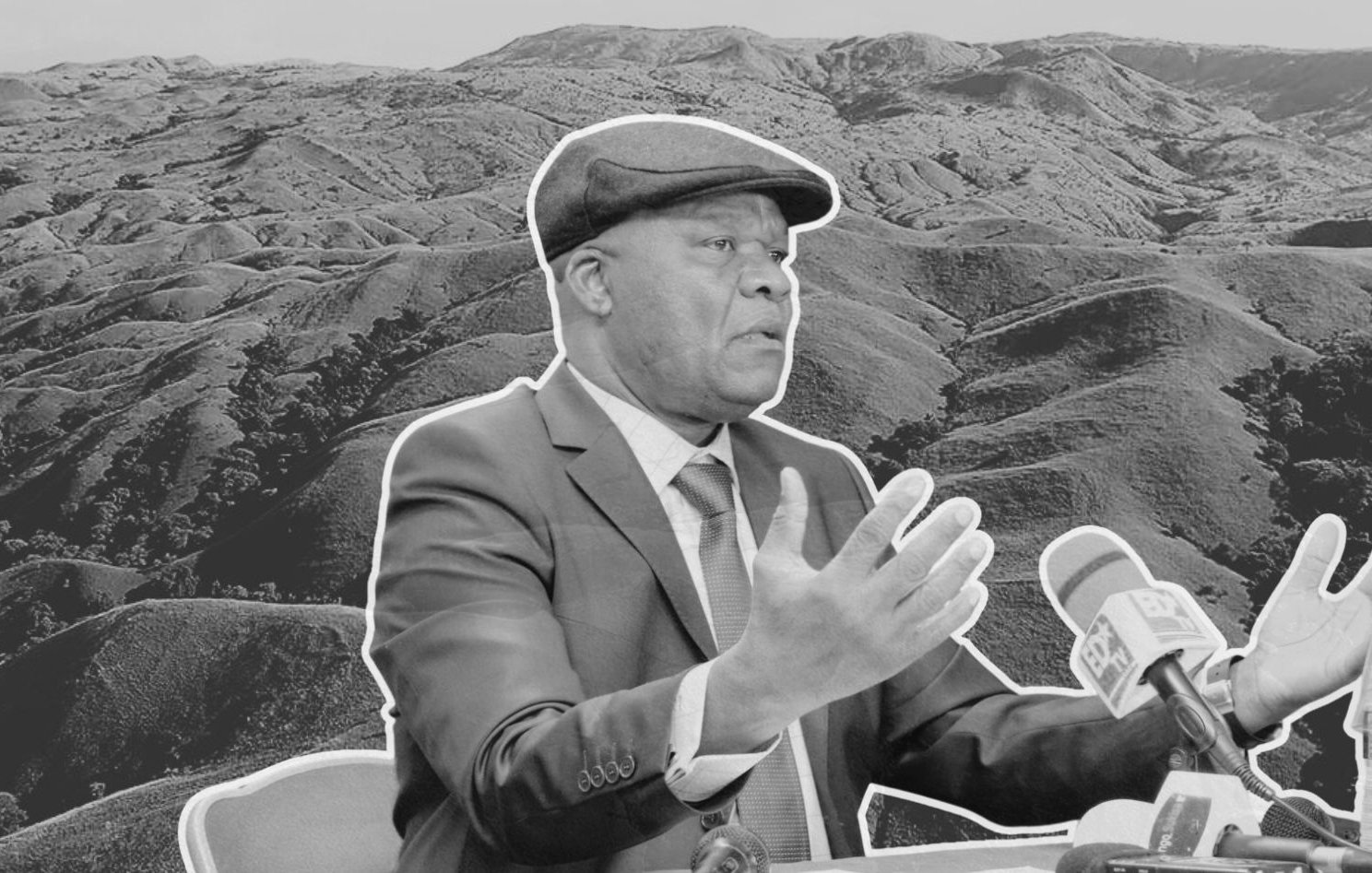 L'image montre un homme en costume, avec un chapeau, en train de parler lors d'une conférence ou d'une interview. Il est assis devant un microphone et semble gesticuler pour appuyer ses propos. En arrière-plan, on aperçoit un paysage de collines ondulantes, évoquant une nature sauvage et montagneuse, donnant une impression de profondeur et de calme. L'ensemble de l'image est en noir et blanc, ce qui renforce le sérieux de la scène.