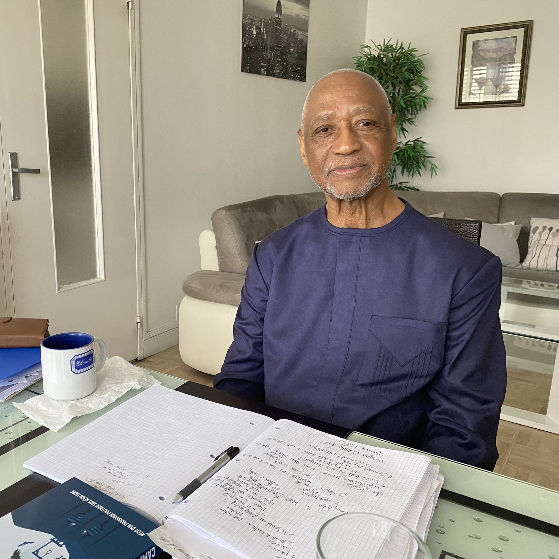 L'image montre un homme assis à une table chez lui. Il porte une tenue traditionnelle de couleur sombre et affiche un sourire chaleureux. La table est en verre et est ornée de divers objets, dont un pot de fleurs vert, un carnet ouvert avec des notes, et un stylo. À côté du carnet, il y a une tasse bleue sur la table. En arrière-plan, on peut voir un canapé clair et des décorations murales. La lumière naturelle entre par une fenêtre, créant une ambiance lumineuse et accueillante.