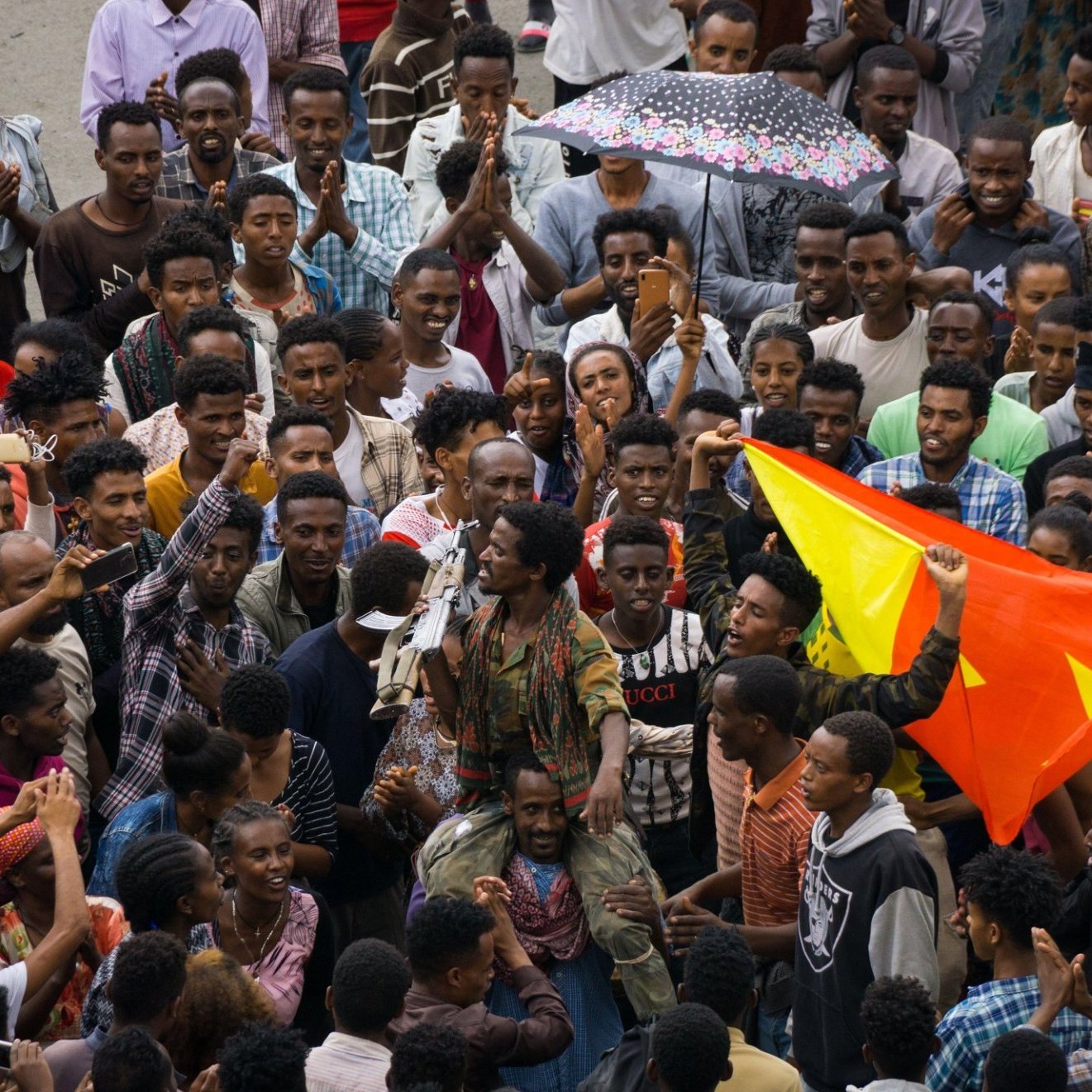 The image depicts a large gathering of people in a vibrant setting, likely representing a celebration or demonstration. Crowds are tightly packed together, with some individuals holding colorful flags, possibly indicating a cultural or political event. One person stands out in the center, engaging the crowd, which looks enthusiastic and expressive. The mood appears lively, with a sense of unity among the participants. Various expressions and clothing styles suggest a diverse group, reflecting a shared purpose or message. An umbrella can be seen, hinting at the weather conditions. Overall, it captures a moment of collective energy and communal spirit.