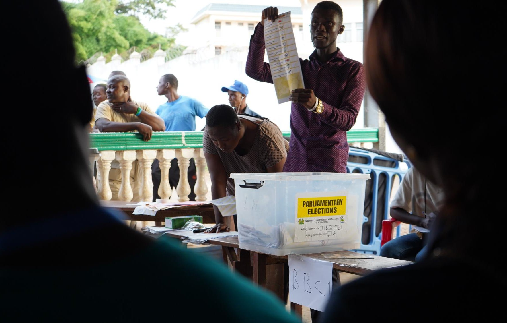 L'image montre une scène d'élections parlementaires. Au centre, un homme se tient debout, levant une feuille de papier, probablement un bulletin de vote, pour montrer aux personnes présentes. Il s'habille d'une chemise de couleur sombre et semble expliquer quelque chose de manière engageante. En arrière-plan, plusieurs personnes sont rassemblées, certaines observant attentivement tandis que d'autres attendent, adossées à une balustrade. On devine une atmosphère de concentration et d'anticipation, typique d'un moment électoral. Sur une table, une boîte transparente étiquetée "ELECTIONS PARLEMENTAIRES" est visible, contenant probablement des bulletins de vote. 