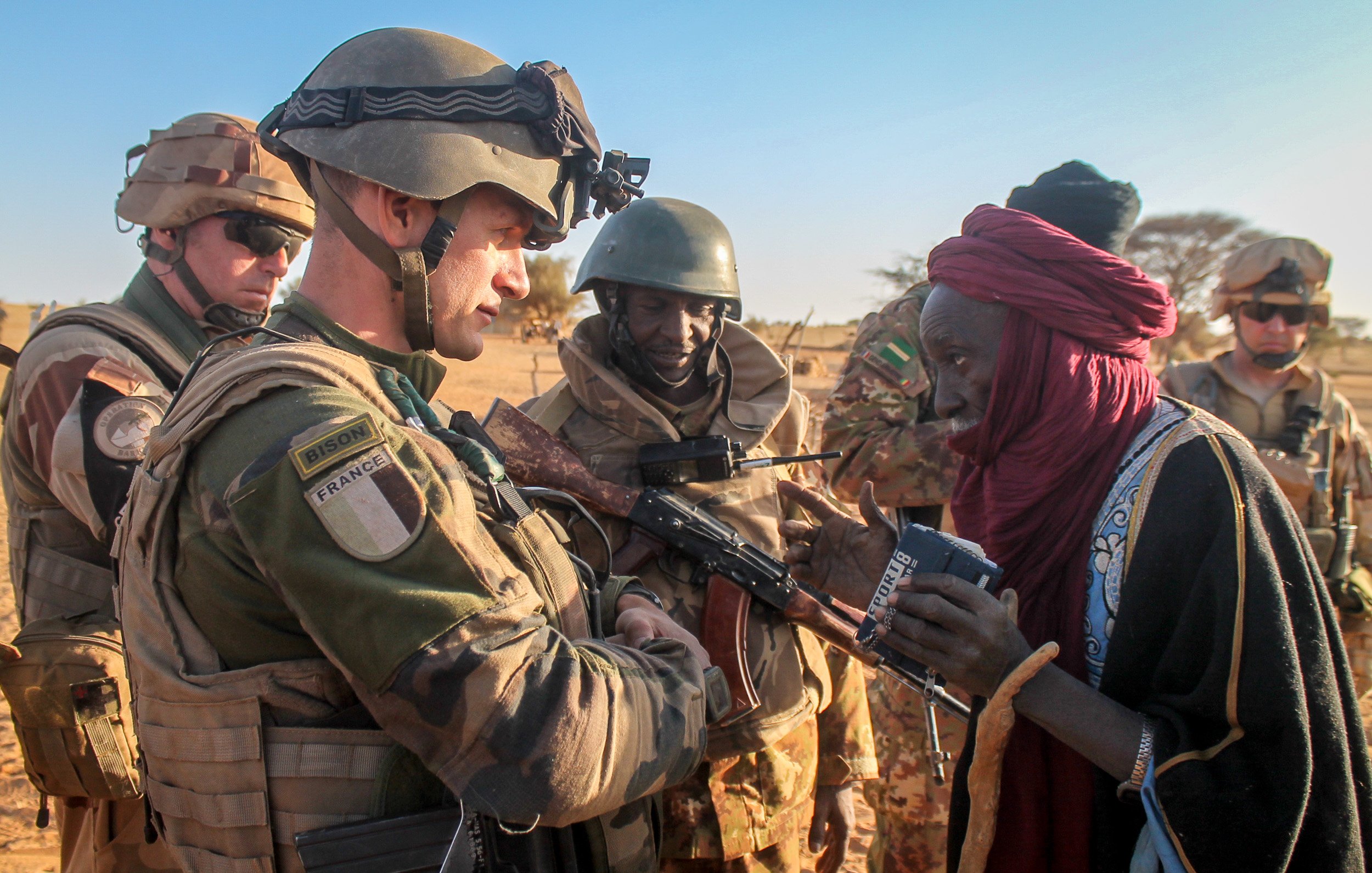 L'image montre une scène dans un environnement désertique, probablement au Sahel. Au premier plan, un soldat en uniforme militaire, portant un gilet pare-balles et un casque, interagit avec une femme vêtue d'une robe traditionnelle. Elle a l'air âgée et semble lui parler tout en tenant un objet dans ses mains. D'autres soldats en arrière-plan, arborant également des uniformes militaires, observent la scène avec un air attentif. L'éclairage est naturel, suggérant que la photo a été prise en fin d'après-midi, et le paysage environnant est sec avec des dunes de sable visibles. Cette rencontre reflète peut-être une interaction humanitaire ou de soutien entre les militaires et la population locale.