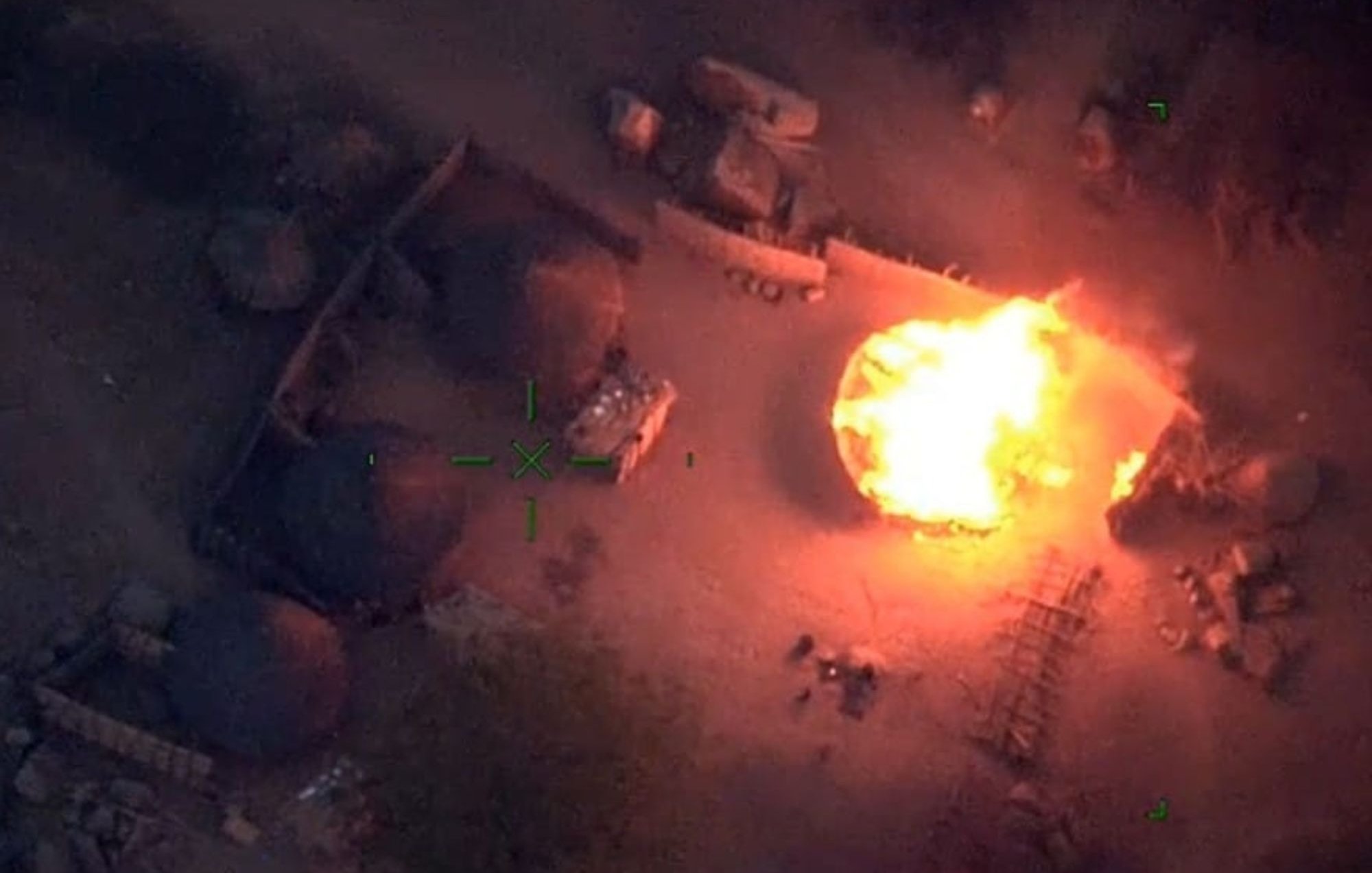 L'image représente une scène vue du ciel, où l'on aperçoit un environnement rural. Au centre, une grande flamme intense monte vers le ciel, créant une illumination vive dans l'obscurité. Entourant cette flamme, on peut distinguer plusieurs structures, probablement des huttes ou des abris, formées de matériaux naturels. Le sol semble légèrement chaotique, avec des débris dispersés et des ombres créées par les objets environnants. L'atmosphère est dramatique et évoque une situation de tension ou de conflit.