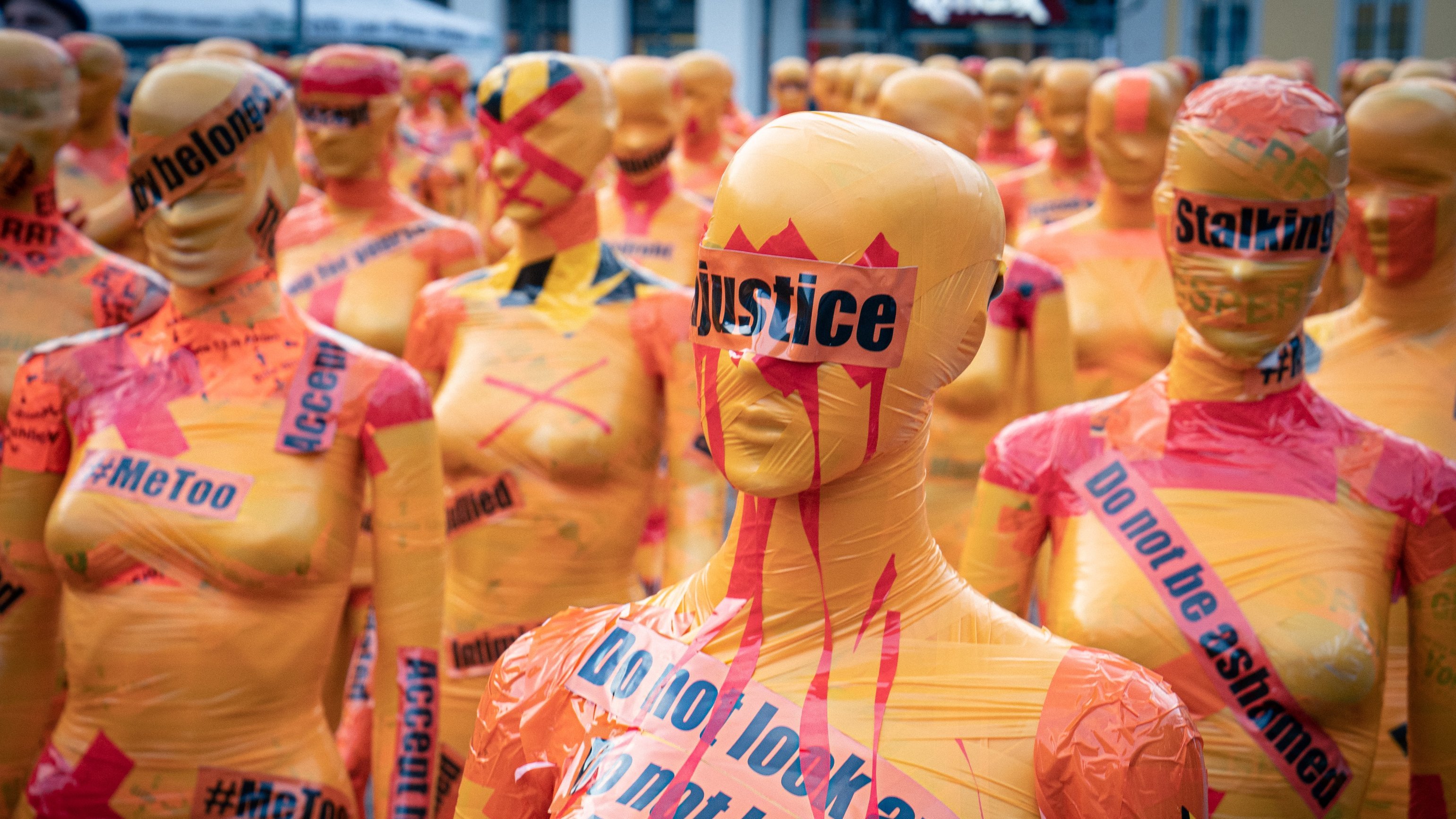 L'image montre un vaste rassemblement de silhouettes humaines couvertes de plastique coloré. Chaque silhouette est peinte en orange vif et ornée de mots et de slogans en noir et en rouge, évoquant des thèmes sociaux et des mouvements de droits civiques comme #MeToo et la justice. Certains mots sont barrés ou soulignés pour accentuer leur impact. L'atmosphère dégage un sentiment fort d'engagement et de protestation, avec ces figures anonymes qui semblent former un mur de revendication. L'ensemble crée une installation artistique saisissante, incitant à la réflexion sur des enjeux sociaux importants.