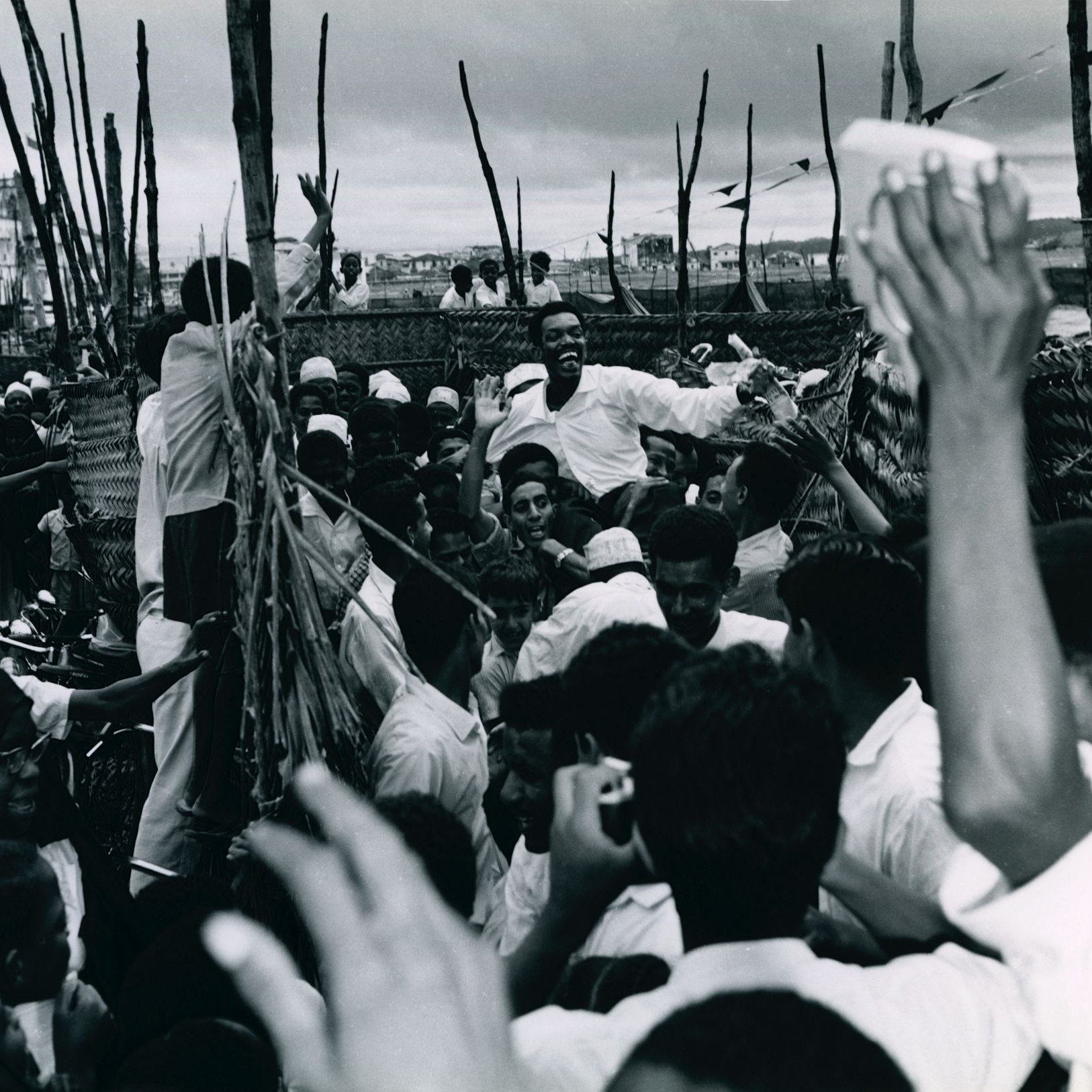L'image montre une scène animée et dynamique, probablement lors d'un rassemblement ou d'une célébration. De nombreuses personnes se regroupent, leurs visages expriment la joie et l'excitation. On peut voir des hommes et des femmes, certains portant des vêtements traditionnels. Au centre, un homme semble chercher à atteindre un groupe, suscitant des réactions enthousiastes de la foule. Les bras de plusieurs personnes s'élèvent, clairement engagés dans l'événement. L'arrière-plan apparaît flou, avec des éléments de construction ou des arbres qui ajoutent à l'atmosphère vivante de la scène. L'ambiance générale évoque une forte connexion et une communauté vibrante.