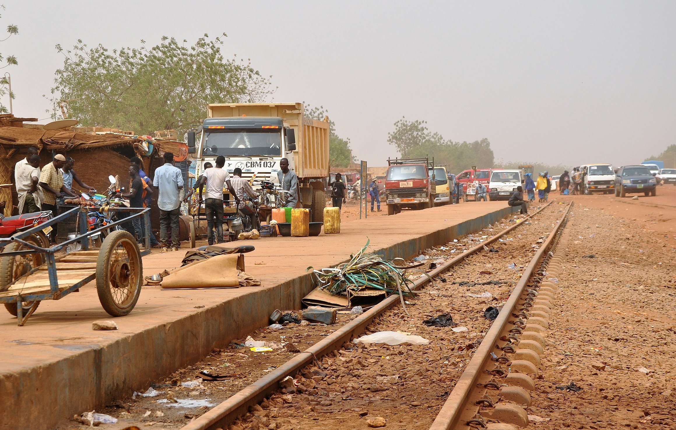 L'image dépeint une scène animée le long d'une voie ferrée. Sur la droite, des rails en métal s'étendent, entourés de poussière. À proximité, on aperçoit plusieurs véhicules, notamment des camions et des voitures, stationnés ou en mouvement. Des personnes se regroupent sur le côté, certains discutant, d'autres transportant des marchandises. Sur le sol, on peut distinguer divers objets laissés là, comme des déchets, des morceaux de métal et des câbles. À gauche, une petite charrette à deux roues est partiellement visible. En arrière-plan, la végétation est clairsemée, avec quelques arbres. Le ciel est d'un ton brumeux, suggérant une chaleur intense et un environnement rural. L'atmosphère générale est à la fois occupée et rustique, évoquant l'activité quotidienne dans une région où la vie s'organise autour des transports.