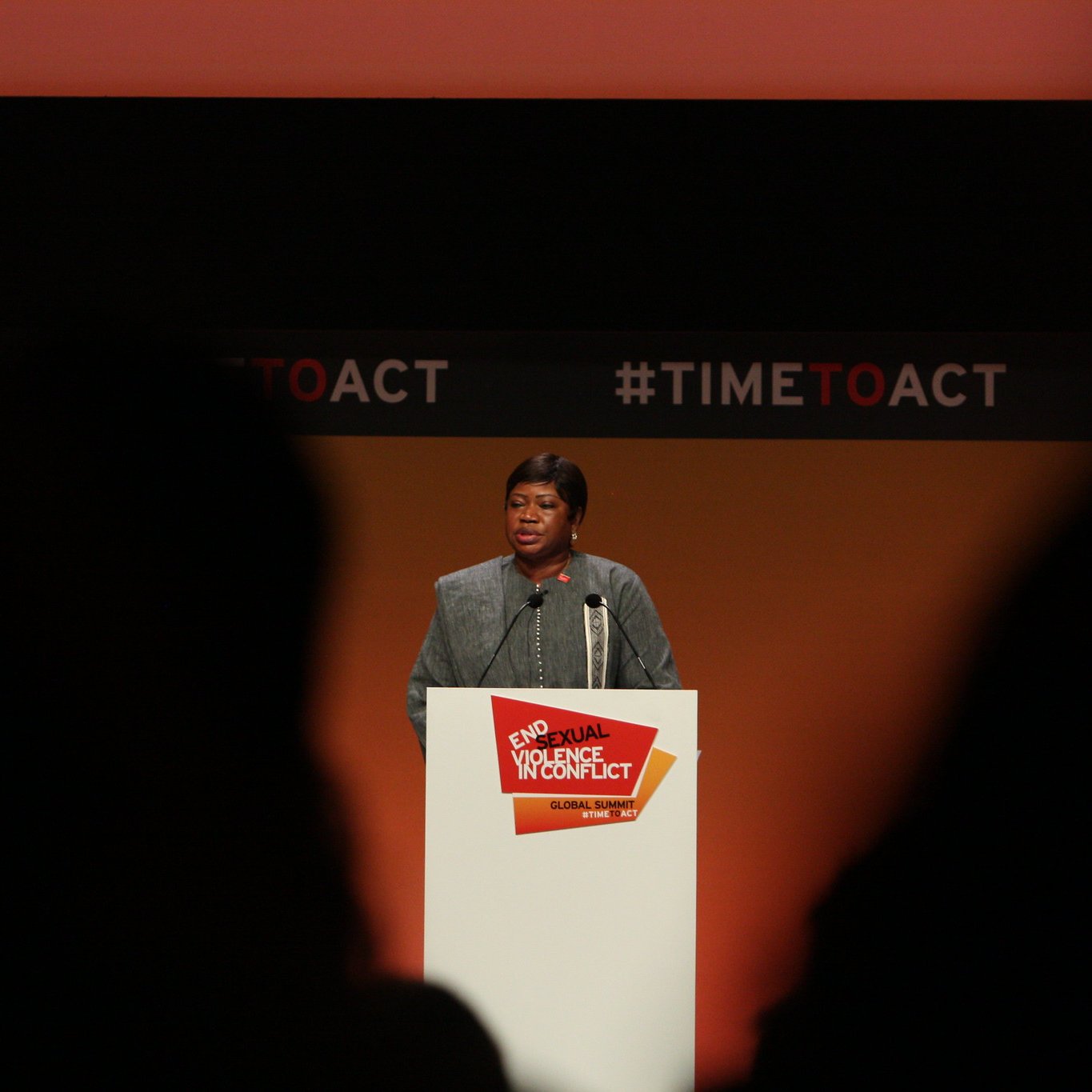 L'image montre une femme debout au micro, s'adressant à un public. Elle porte un vêtement formel et a une expression sérieuse. Le fond est principalement orange, avec des éléments graphiques en noir et rouge. Un panneau blanc devant elle affiche le texte "End Sexual Violence in Conflict" et "#TIMETOACT". La scène semble être un événement ou une conférence sur un sujet important, probablement lié à la lutte contre la violence sexuelle en contexte de conflit. Les silhouettes des personnes dans le public sont visibles en bas de l'image, ajoutant à l'ambiance de l'événement.