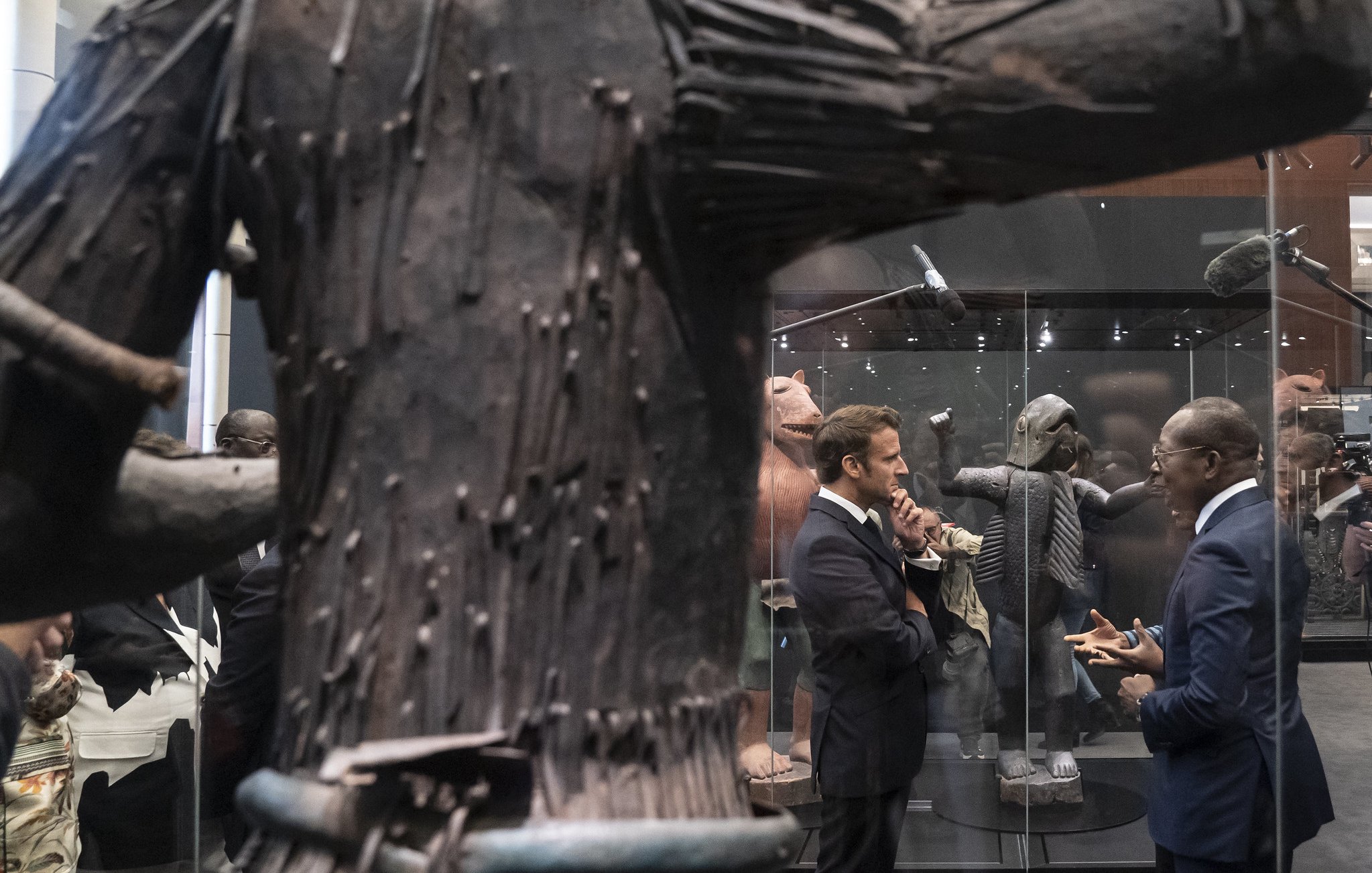 L'image montre un espace d'exposition où deux hommes en costume discutent. L'un d'eux semble concentré sur la conversation, tandis que l'autre écoute attentivement. Ils se tiennent près d'une grande sculpture en bois, qui occupe une place centrale. Cette sculpture a une texture rugueuse, avec des détails qui évoquent une forme humaine ou abstraite. En arrière-plan, d'autres œuvres d'art sont visibles, ajoutant à l'atmosphère culturelle de l'exposition. L'éclairage est doux, mettant en valeur les sculptures tout en créant une ambiance contemplative et respectueuse.