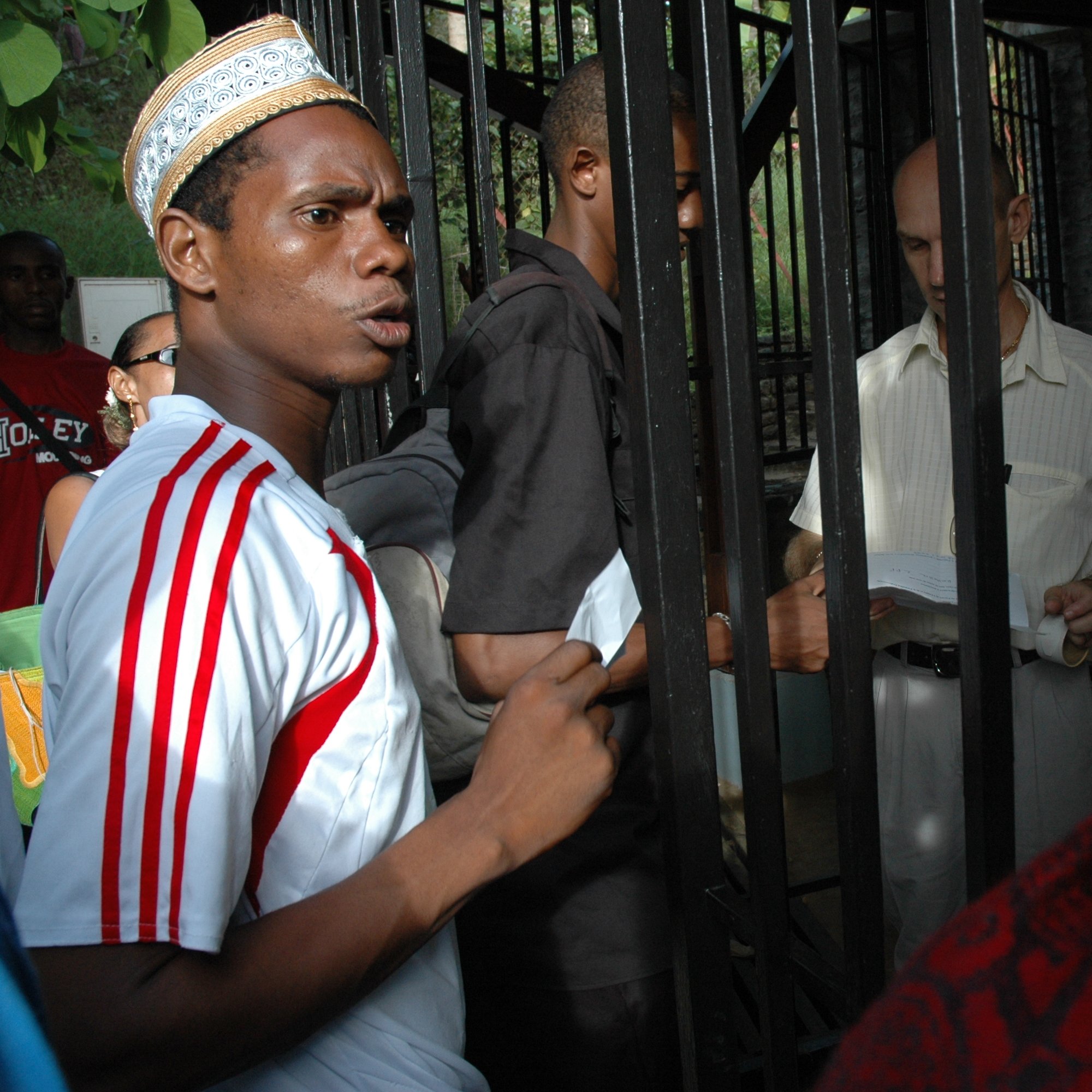 L'image montre un groupe de personnes rassemblées autour d'une structure métallique, probablement une grille ou une clôture. Un jeune homme au premier plan, portant un maillot blanc avec des rayures rouges et un chapeau traditionnel, exprime une certaine agitation ou curiosité. À côté de lui, d'autres personnes, dont un homme en chemise claire, semblent discuter ou échanger des documents. L'environnement est extérieur et verdoyant, suggérant un lieu fréquenté. Les expressions des visages indiquent une interaction dynamique et engageante entre les personnes présentes.
