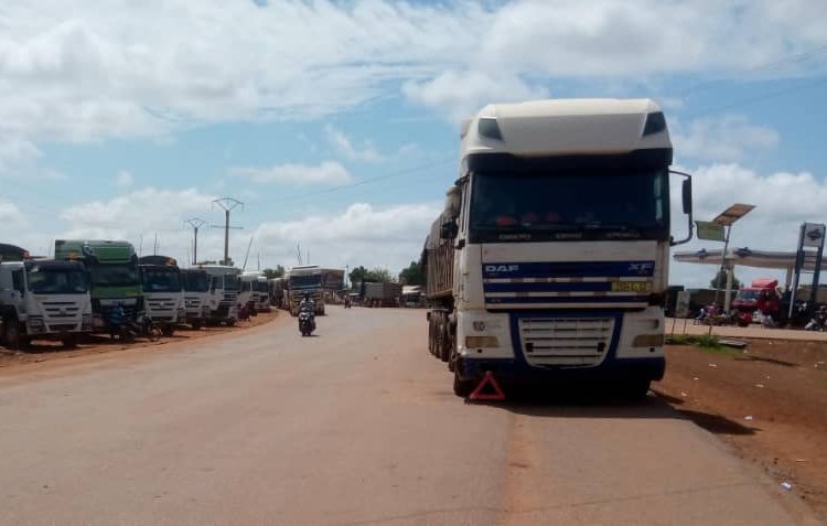 L'image montre une route large et droite, où une file de camions est stationnée sur le côté. Les camions, de différentes tailles et couleurs, sont alignés de manière ordonnée. Le ciel est clair avec quelques nuages blancs qui flottent. On peut apercevoir des poteaux électriques le long de la route, et au loin, il y a des bâtiments, suggérant un environnement urbain en arrière-plan. À proximité, un motocycliste passe, ajoutant un peu de dynamisme à la scène calme. L'ambiance générale dégage une impression de circulation routière et d'activités commerciales.