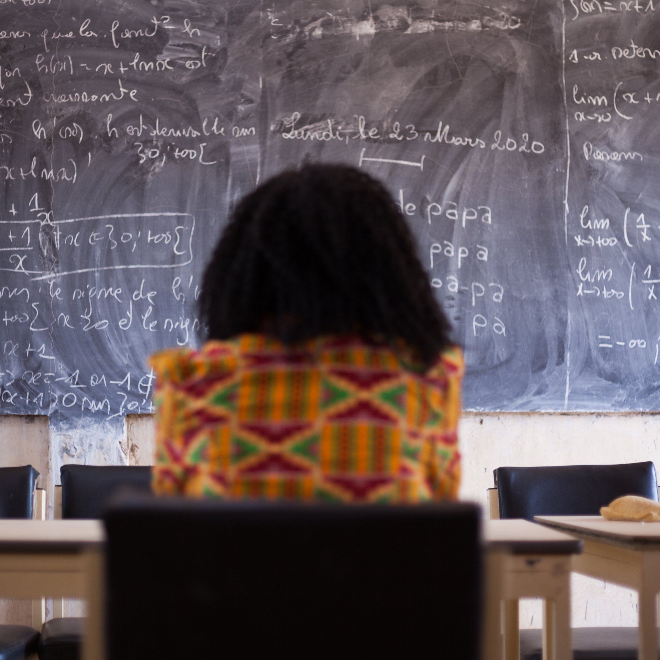 Dans cette image, on voit une personne assise, de dos, face à un grand tableau noir couvert d'écritures en craie blanche. Les formules mathématiques et les notes semblent complexes, suggérant un environnement académique ou d'apprentissage. La personne porte un vêtement coloré avec des motifs vibrants, ajoutant une touche de culture. L'ambiance est studieuse, et l'environnement évoque une salle de classe ou un espace de travail. Le contraste entre les couleurs vives du vêtement et la sombre surface du tableau crée une composition visuelle intéressante.