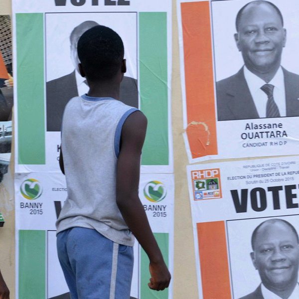 L'image montre un groupe de jeunes garçons marchant le long d'un mur recouvert d'affiches électorales. Les affiches présentent des portraits de candidats politiques, dont certaines affichent le nom d'Alassane Ouattara. Les affiches sont aux couleurs vives, avec des fonds colorés, principalement du rose et du vert, et des mots comme "VOTEZ". Les garçons, vêtus de t-shirts, semblent attentifs aux affiches. Le décor est urbain et évoque l'ambiance d'une campagne électorale animée.