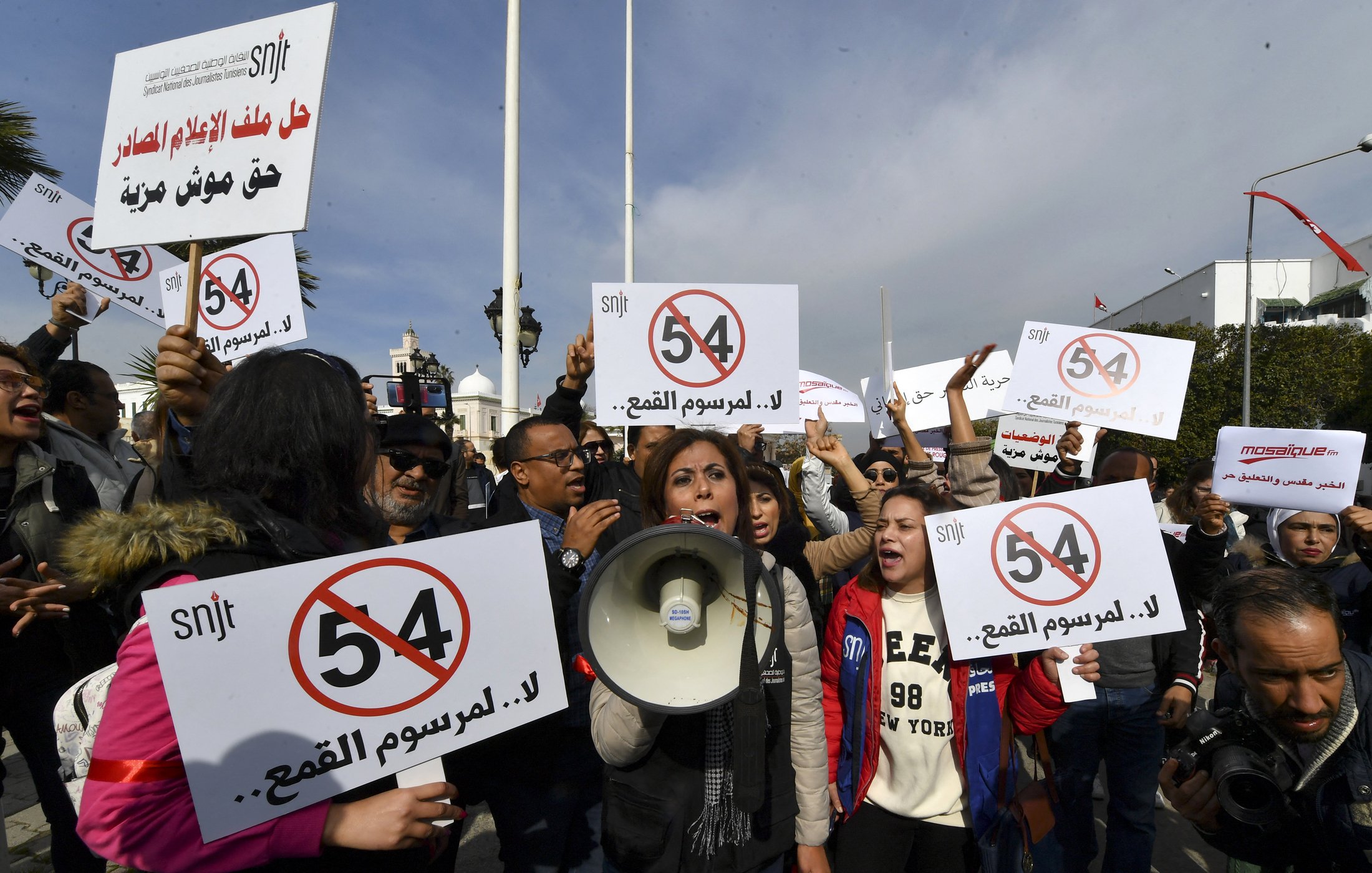 The image depicts a protest scene with a group of people holding signs and banners. In the foreground, a woman is speaking into a megaphone, likely leading the demonstration. The signs feature the number "54" and messages opposing censorship, suggesting that the protest is focused on issues related to freedom of expression or press freedom. The crowd appears to be actively engaged in the protest, raising their signs and voicing their demands. The setting seems to be outdoors, with flags and possibly a government building in the background.