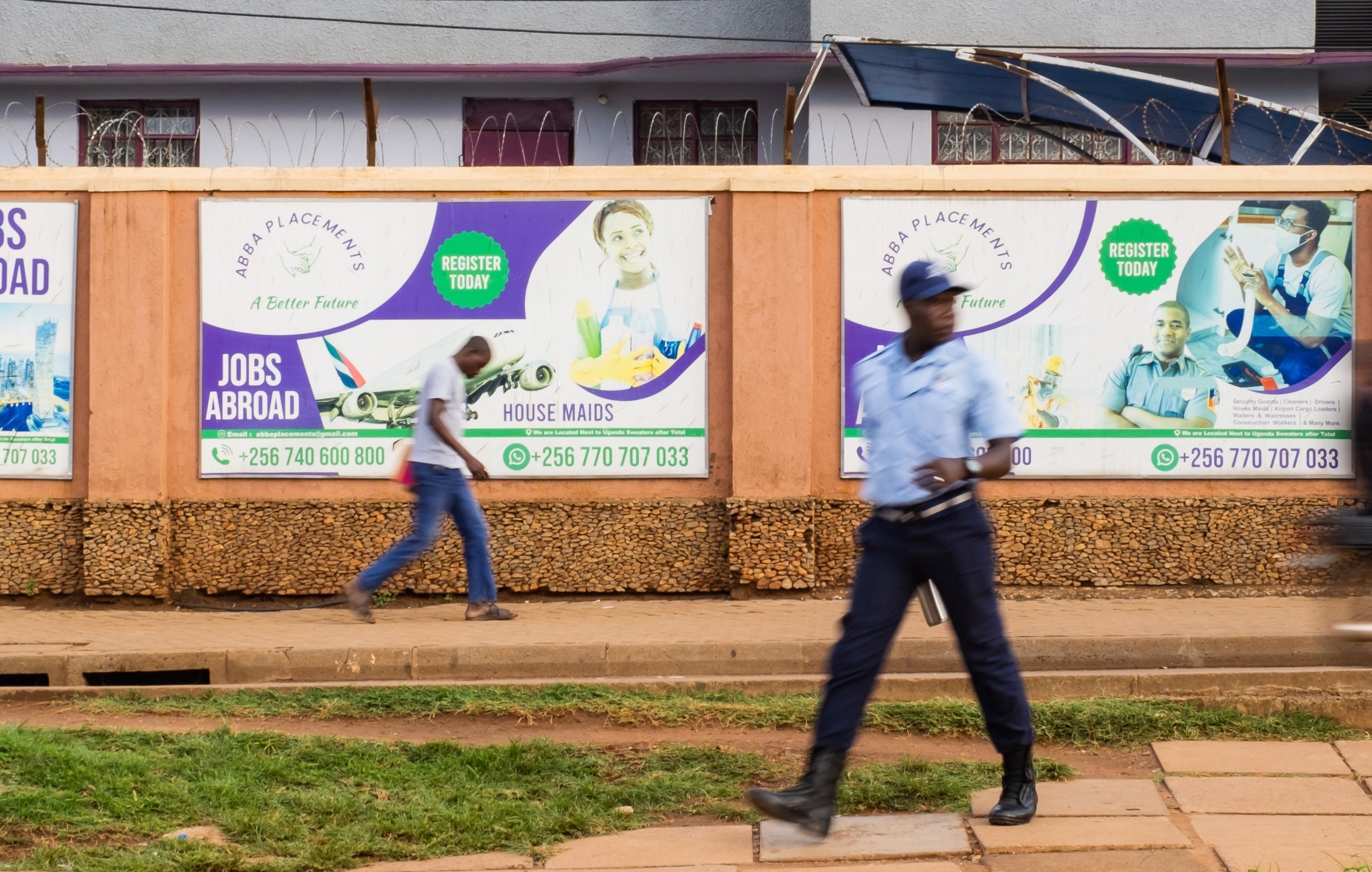 L'image montre un environnement urbain animé. Sur un mur en bois, plusieurs affiches colorées sont accrochées, promouvant des services tels que des opportunités de travail à l'étranger et des services de ménage. Les affiches présentent des visages souriants de personnes, évoquant une atmosphère positive et encourageante. Au premier plan, un homme en uniforme, probablement un agent de sécurité, marche avec assurance sur le trottoir, tandis qu'un autre passant se déplace rapidement derrière lui. Le sol est pavé, et on peut deviner une verdure légère avec une pelouse bien entretenue. L'ensemble évoque une scène de la vie quotidienne dans une ville dynamique.