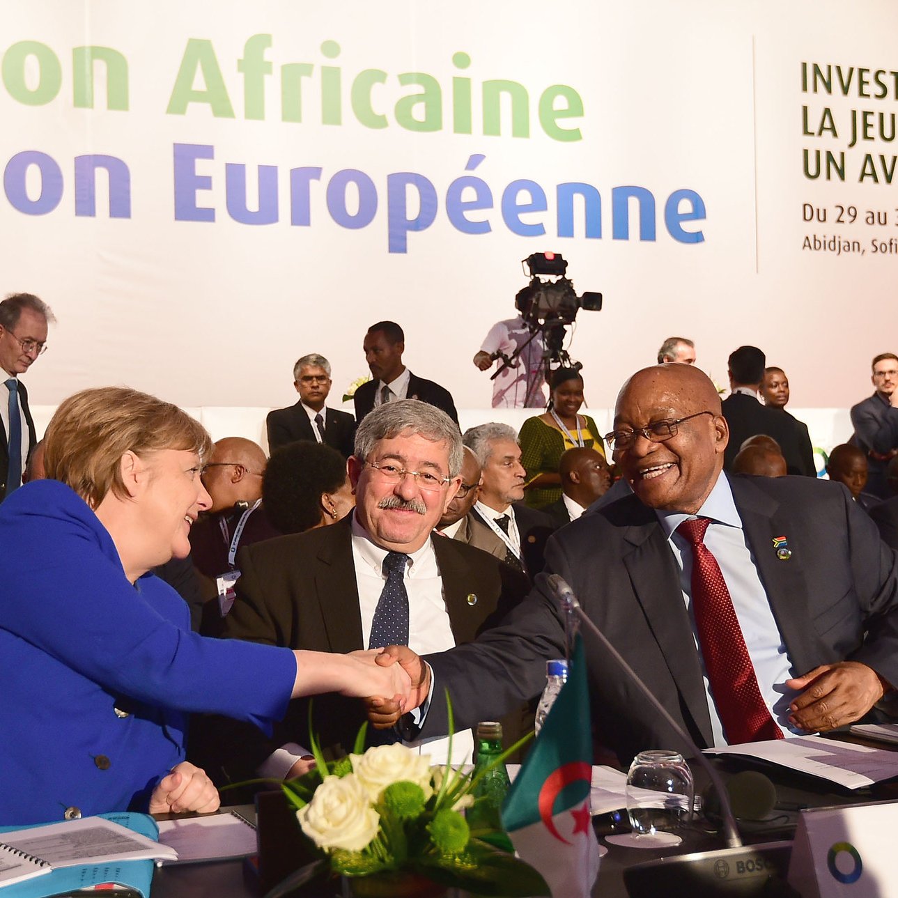 L'image montre la scène d'une conférence internationale où des dirigeants participent à un sommet. Au premier plan, on aperçoit un groupe de personnes, parmi lesquelles une femme, identifiée comme une figure politique importante, est en train de serrer la main d'un homme. Cette interaction symbolise le dialogue et la collaboration entre les nations. À l'arrière-plan, on distingue des bannières et des affiches portant le titre "Union Africaine, Union Européenne" ainsi que le slogan "Investir dans la jeunesse pour un avenir durable". La salle semble remplie de délégués, en costard et concentrés sur les discussions, ce qui donne une atmosphère formelle et engagée. Des éléments comme une table avec des drapeaux de différents pays ajoutent au cadre diplomatique de l'événement.