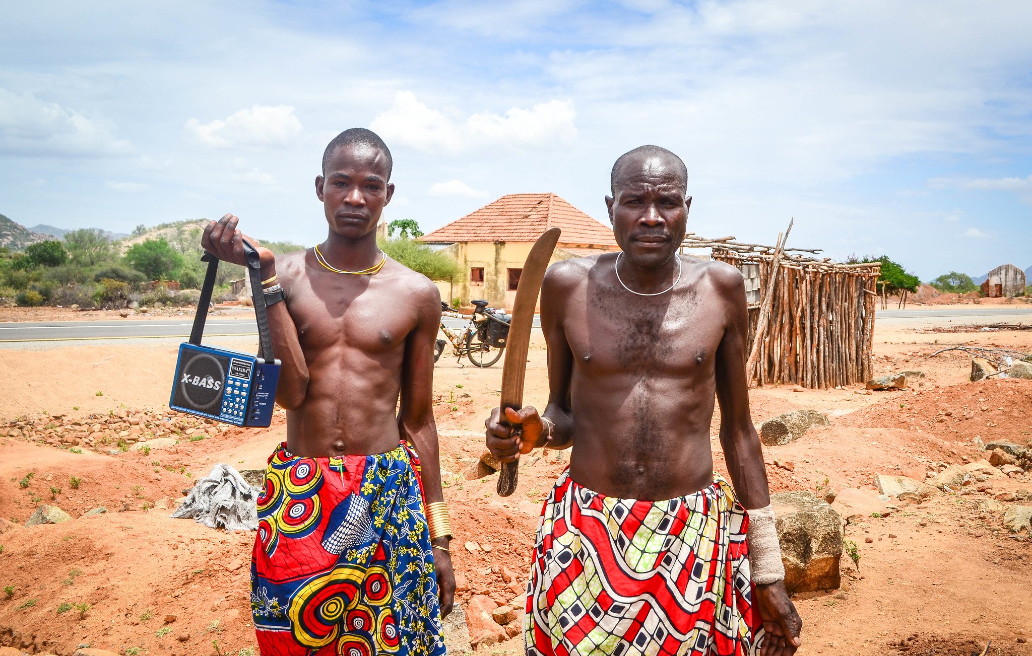 L'image montre deux hommes debout sur un terrain sec et rocheux. Ils portent des vêtements colorés avec des motifs vibrants, typiques d'une culture africaine. Le premier homme, à gauche, tient un poste de radio portable, tandis que le second, à droite, brandit une grande machette. En arrière-plan, on peut apercevoir des bâtiments simples, ainsi qu'une nature aride avec quelques arbres. Le ciel est bleu avec quelques nuages. L'atmosphère semble calme, mais la présence des objets et des vêtements indique une culture dynamique et vivante.