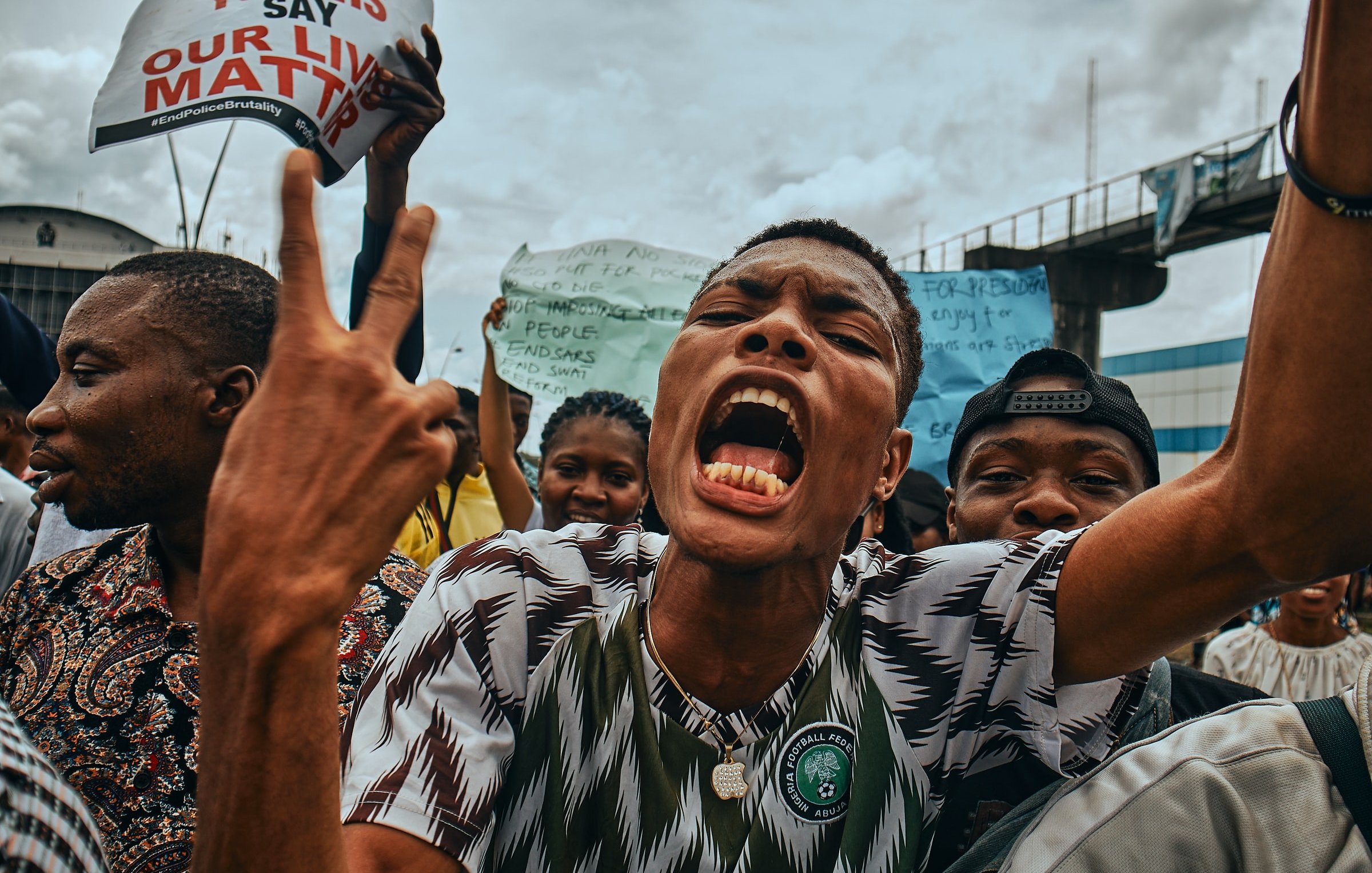 L'image montre une scène de manifestation animée. Au premier plan, un jeune homme crie avec passion, levant les deux doigts en signe de victoire ou de protestation. Son visage exprime une forte intensité émotionnelle, avec une bouche ouverte et des yeux déterminés. Il porte un maillot à motifs verts et blancs, représentant probablement les couleurs de son pays. Derrière lui, un groupe de personnes brandit des pancartes avec des slogans, illustrant un sentiment de mobilisation et de solidarité. L'ambiance est vibrante, évoquant une lutte collective pour des causes qui leur tiennent à cœur, sous un ciel nuageux.