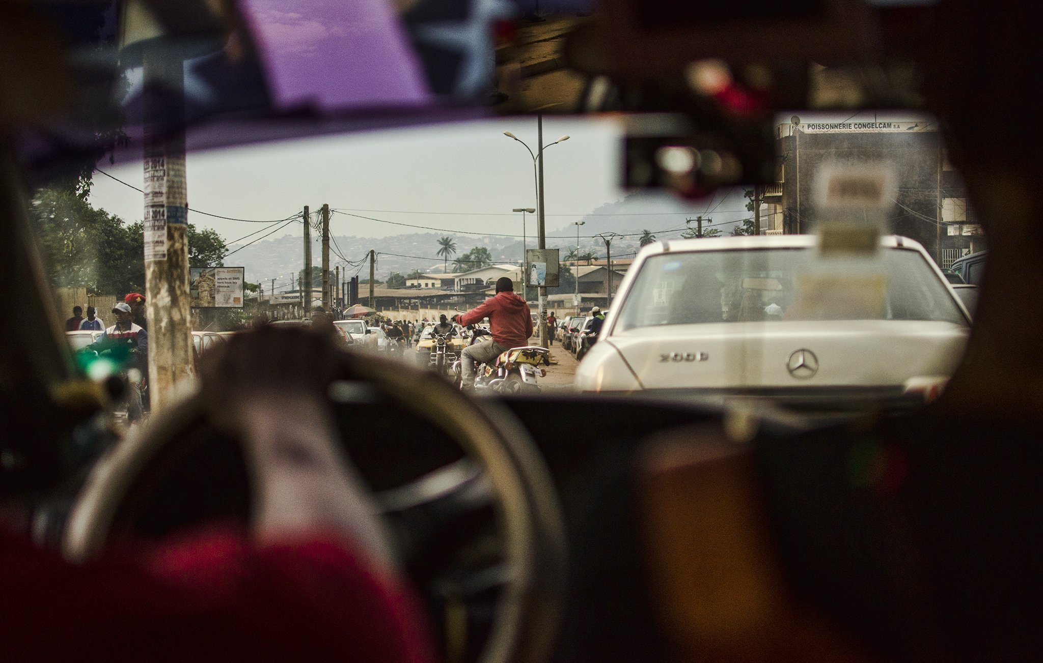 L'image présente une scène urbaine animée vue depuis l'intérieur d'un véhicule. On peut voir le côté d'un tableau de bord, avec une main tenant le volant. À travers le pare-brise, la circulation est dense, comprenant des voitures et des motos. On distingue une personne à vélo sur la route, et au fond, des bâtiments et des montagnes se dessinent à l'horizon. L'atmosphère semble vivante et mouvementée, caractéristique de la vie citadine. La lumière du jour éclaire la scène, révélant des détails sur les véhicules et les personnes qui circulent.