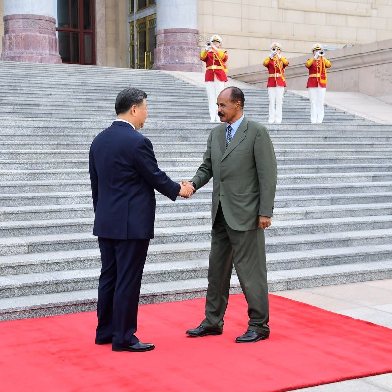 L'image montre deux hommes se serrant la main sur un tapis rouge, symbolisant un accueil officiel. Ils sont debout devant un bâtiment imposant, avec des marches qui mènent à l'entrée. En arrière-plan, on aperçoit des gardes en uniformes, debout et formant une ligne. Le premier homme porte un costume sombre, tandis que le second est habillé d'un costume vert. L'atmosphère dégage un sentiment de cérémonie et de respect mutuel.
