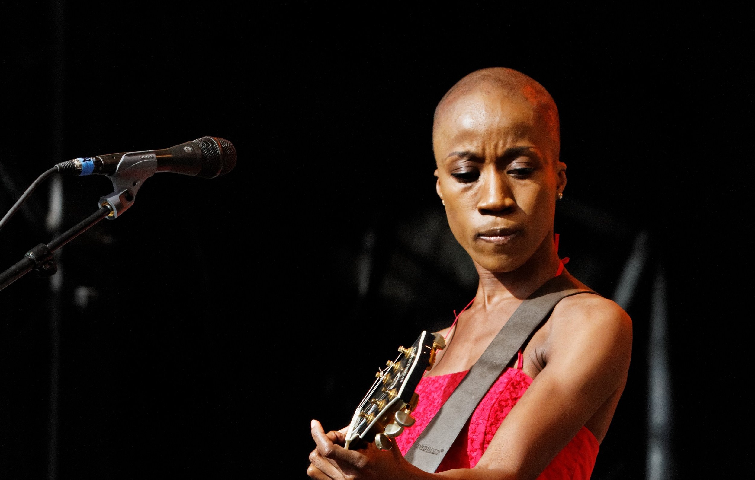 L'image montre une artiste sur scène qui joue de la guitare. Elle a la tête rasée, ce qui met en avant son visage expressif. Elle porte une robe rouge qui attire l'attention et contraste avec l'éclairage du fond sombre de la scène. Son regard est concentré, montrant sa passion et son engagement dans sa performance. Le microphone est placé devant elle, indiquant qu'elle chante probablement en même temps qu'elle joue. L'ambiance générale semble dynamique et remplie de musique, créant une atmosphère captivante.