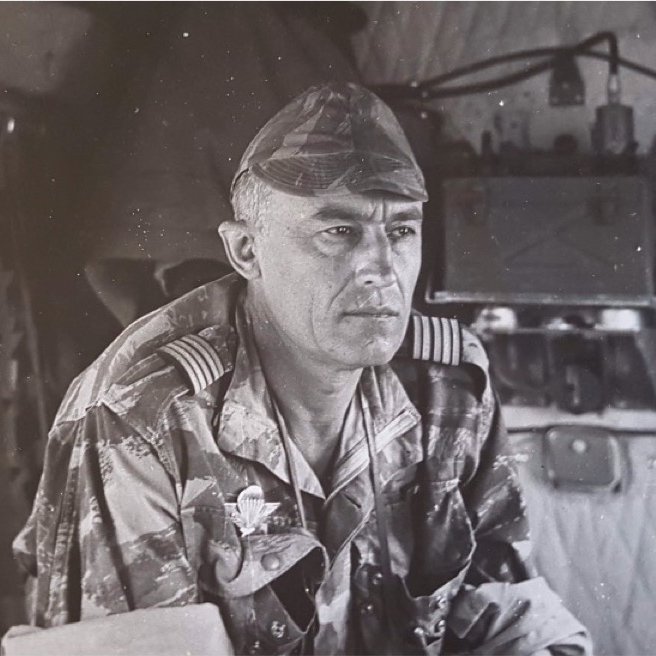 L'image présente un homme en uniforme militaire, assis à l'intérieur d'un véhicule. Il a les cheveux courts et grisonnants, avec une expression sérieuse et réfléchie. Son uniforme, camouflé, est orné d'insignes distinctifs sur les épaules, indiquant son grade. Au fond, on aperçoit des équipements militaires et des matériaux qui suggèrent un environnement de travail opérationnel. La lumière semble douce, et l'atmosphère générale dégage un sentiment de concentration et de détermination.