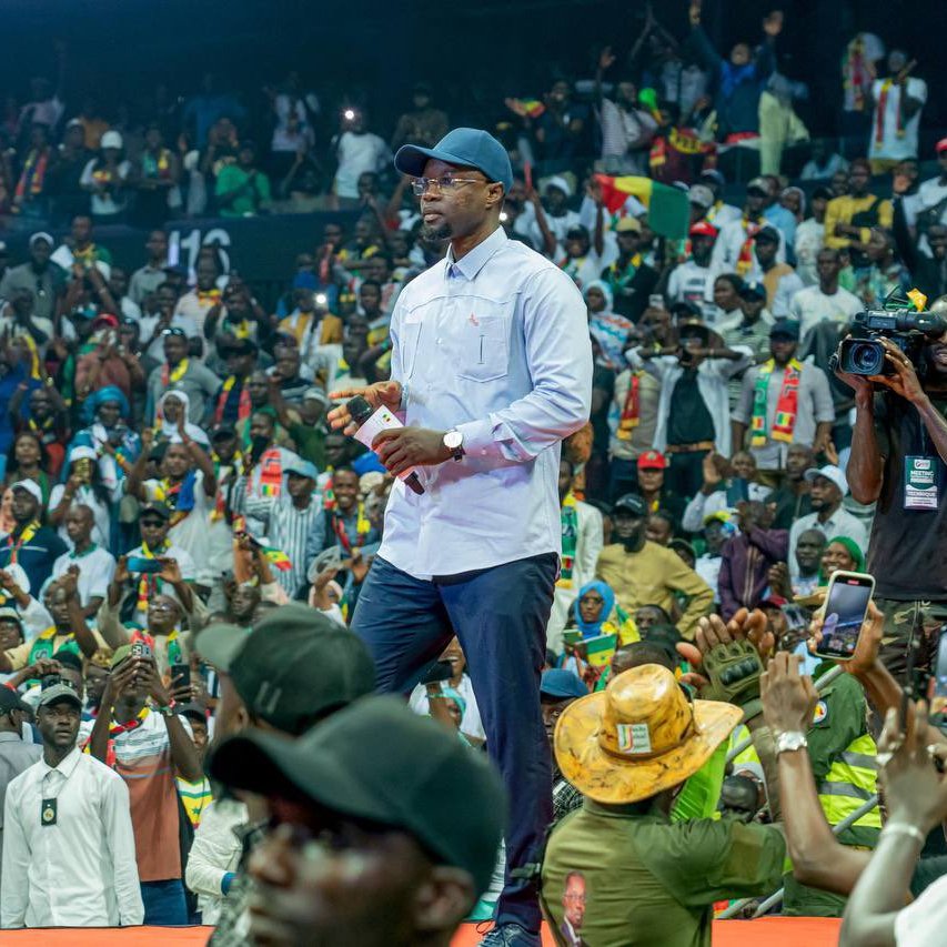 L'image représente un événement animé dans une grande salle remplie de personnes. Au premier plan, un homme en tenue décontractée, portant une chemise claire et une casquette, marche avec assurance sur la scène. Il semble interagir avec le public, qui se compose de nombreux spectateurs enthousiastes, certains levant les mains en signe d'encouragement. La foule est colorée, avec des personnes habillées en diverses tenues, et des drapeaux ou accessoires visibles, suggérant un fort esprit communautaire. En arrière-plan, on peut voir des caméras et des membres de l'équipe de sécurité, ce qui indique que l'événement est de grande envergure. L'atmosphère est d'excitation et d'engagement collectif.