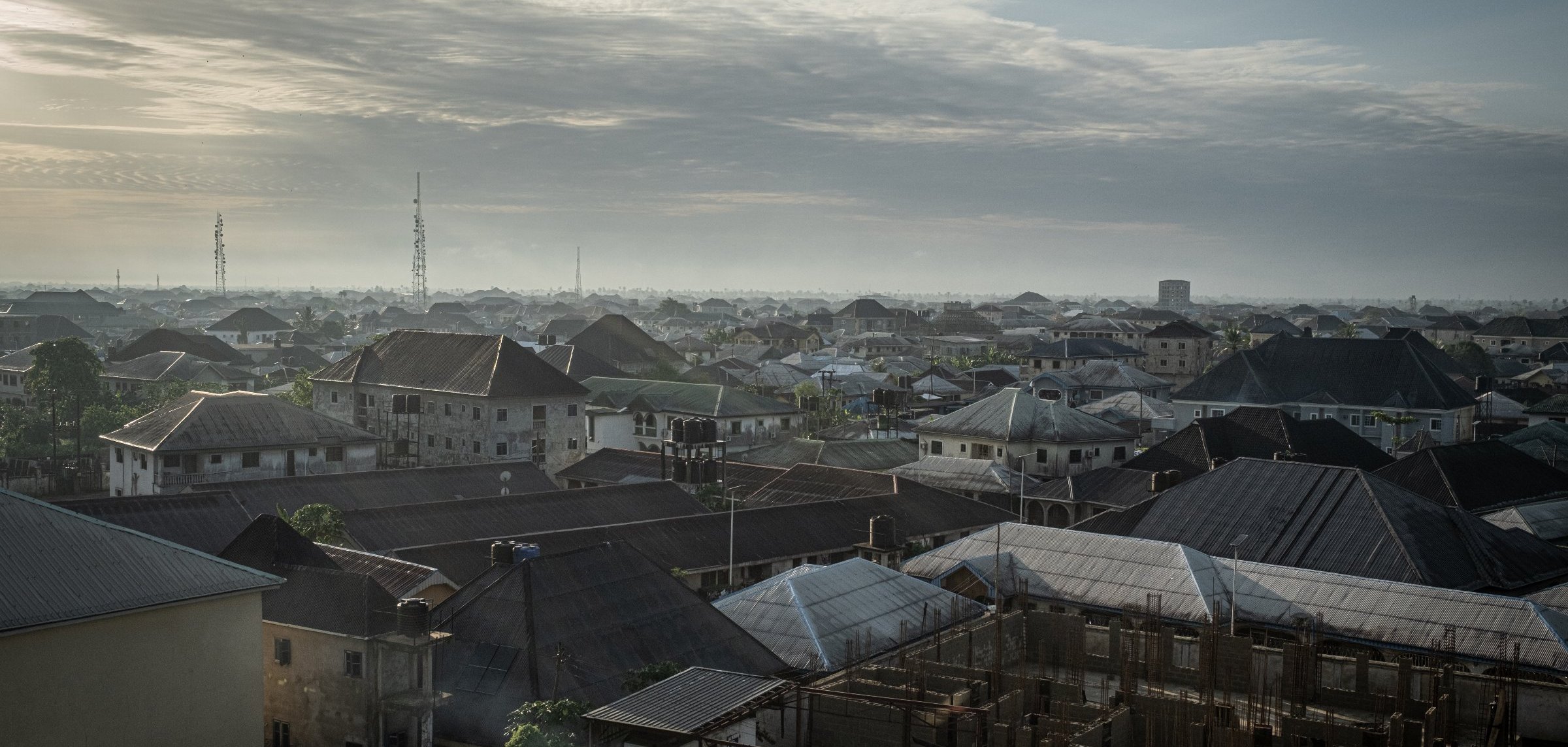 Dans cette image, on voit un paysage urbain d'une ville d'Afrique, probablement au lever du soleil. Les toits de différentes maisons se chevauchent, formant une mosaïque de formes et de textures. La majorité des toits sont en tôle, certains ayant des angles abrupts, tandis que d'autres sont plats. En arrière-plan, on aperçoit des constructions en cours, avec des structures en béton qui indiquent un développement urbain. Le ciel est parsemé de nuages, offrant une lumière douce et diffuse. On devine une atmosphère paisible au matin, avec des ombres légères qui accentuent les contours des bâtiments. Au loin, on distingue des antennes et des colonnes, signes de l'urbanisation moderne. L'ensemble dégage une impression de vie et d'activité, mais aussi de tranquillité à cette heure matinale.