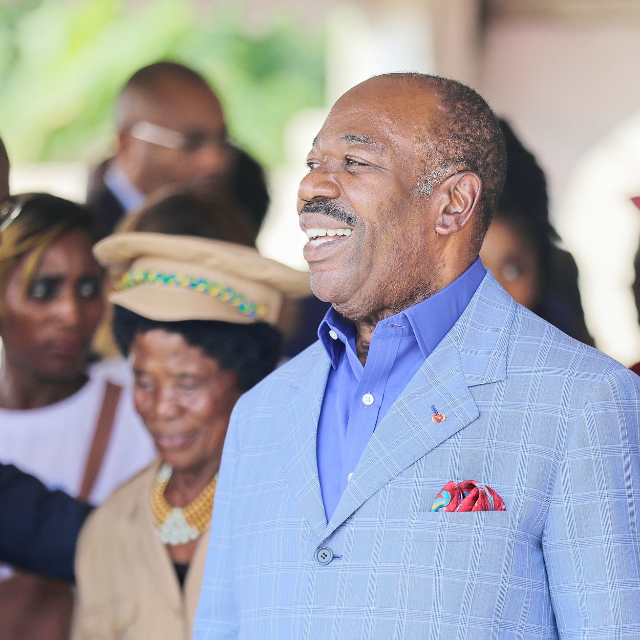L'image montre un groupe de personnes en train d'interagir dans un cadre convivial. Au centre, un homme, portant un costume bleu clair et une chemise bleue, sourit chaleureusement, affichant une attitude amicale et accessible. On note également des personnes en arrière-plan, certaines portant des vêtements colorés, qui semblent engagées dans une conversation ou une activité sociale. L'ambiance globale de l'image est joyeuse et accueillante, suggérant un moment de célébration ou de rencontre.