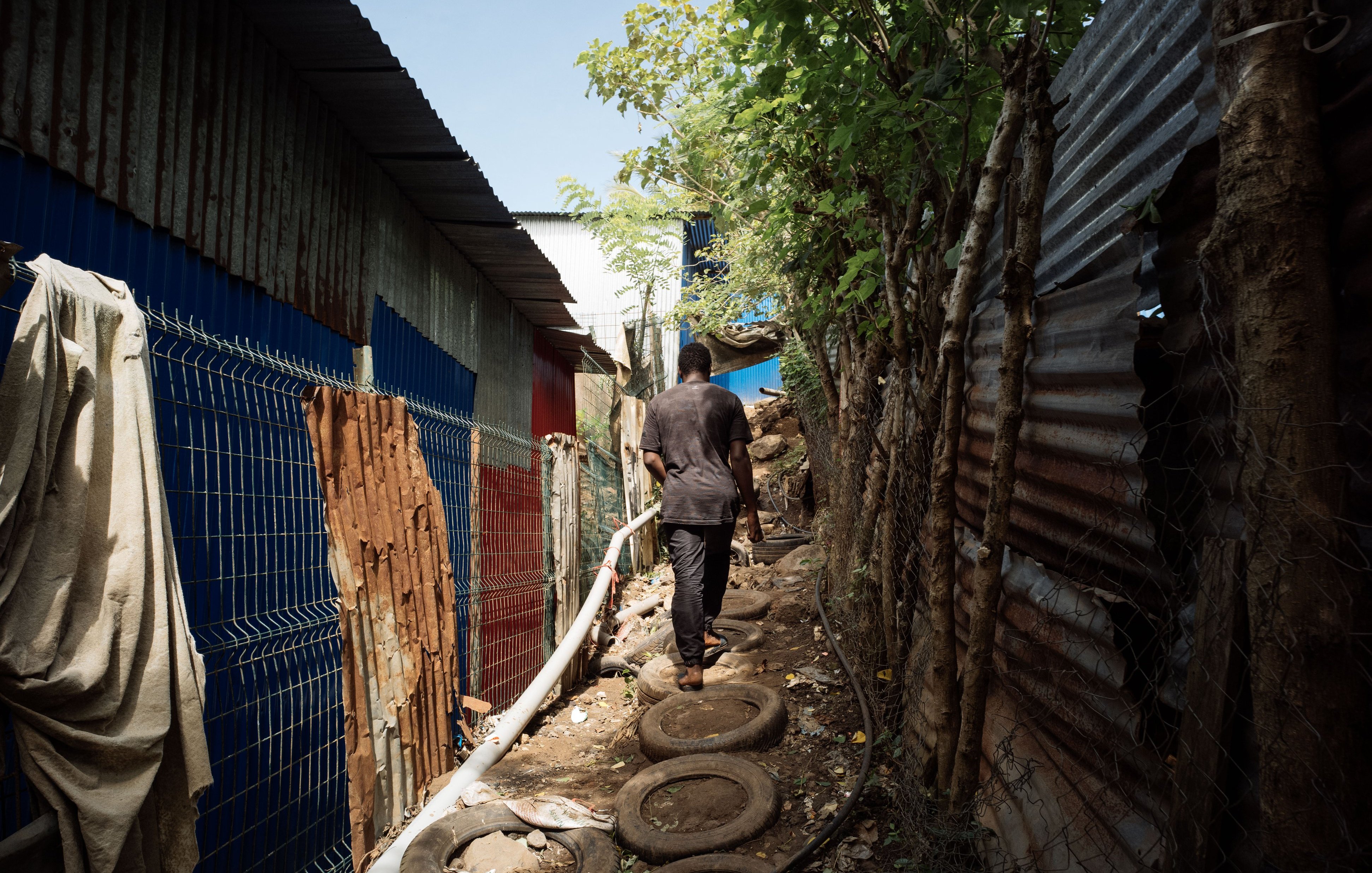 L'image montre un sentier étroit entre deux structures en tôle ondulée, typiques des bidonvilles. À gauche, on distingue des murs colorés, tandis qu'à droite, la structure est recouverte de tôles rouillées. Le chemin est constitué de pneus empilés, et un homme marche le long de ce chemin, portant des vêtements sombres. La lumière du jour illumine la scène, mettant en évidence la végétation qui borde le sentier, avec des arbres et des feuilles qui apportent une touche de verdure à cet environnement urbain. L'atmosphère semble à la fois animée et paisible.