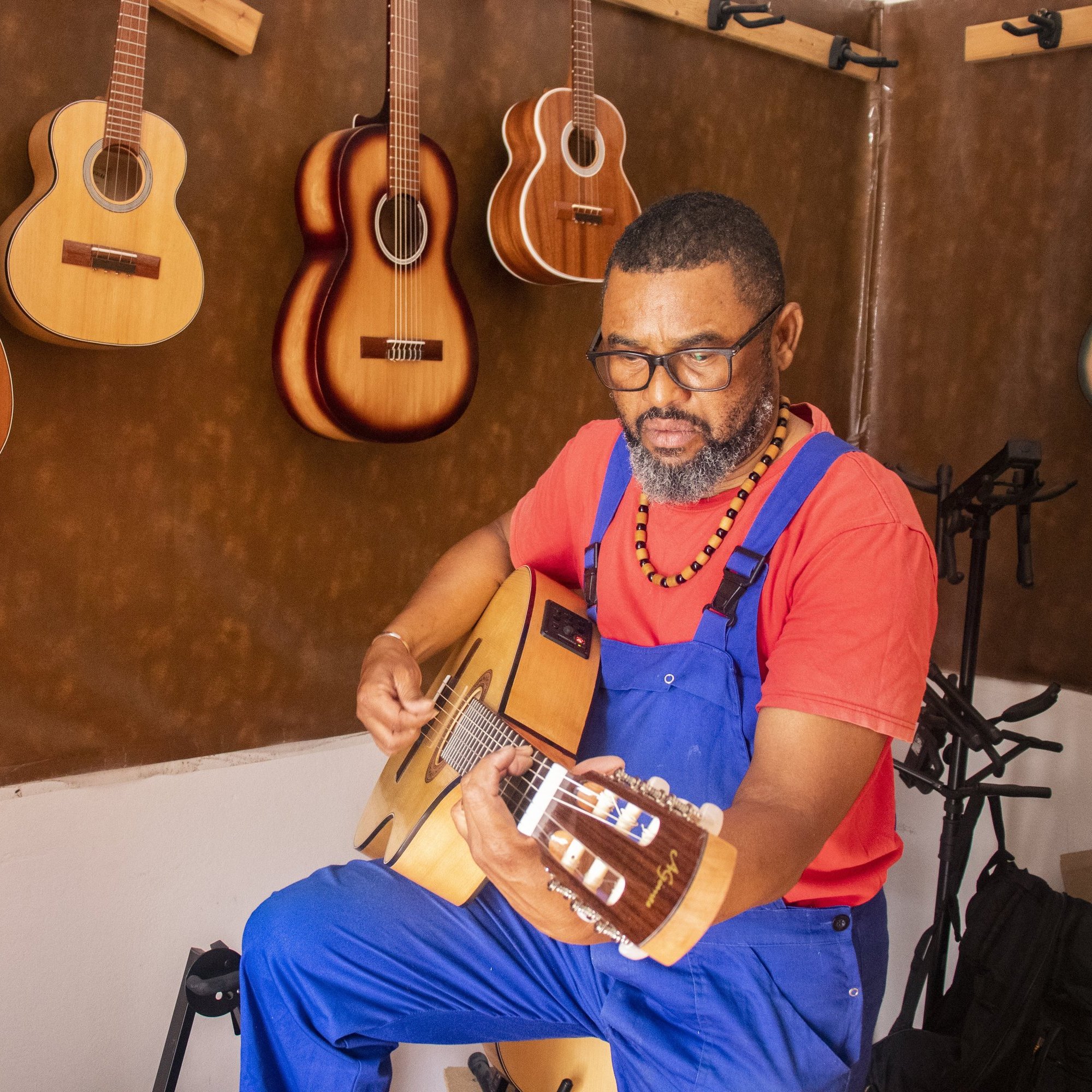 L'image montre un homme assis, vêtu d'une salopette bleue et d'un t-shirt rouge. Il est en train de jouer de la guitare acoustique avec une expression de concentration sur son visage. Autour de lui, plusieurs guitares sont accrochées au mur, dans des teintes de bois naturel. L'ambiance de l'espace est chaleureuse et artistique, soulignant sa passion pour la musique.