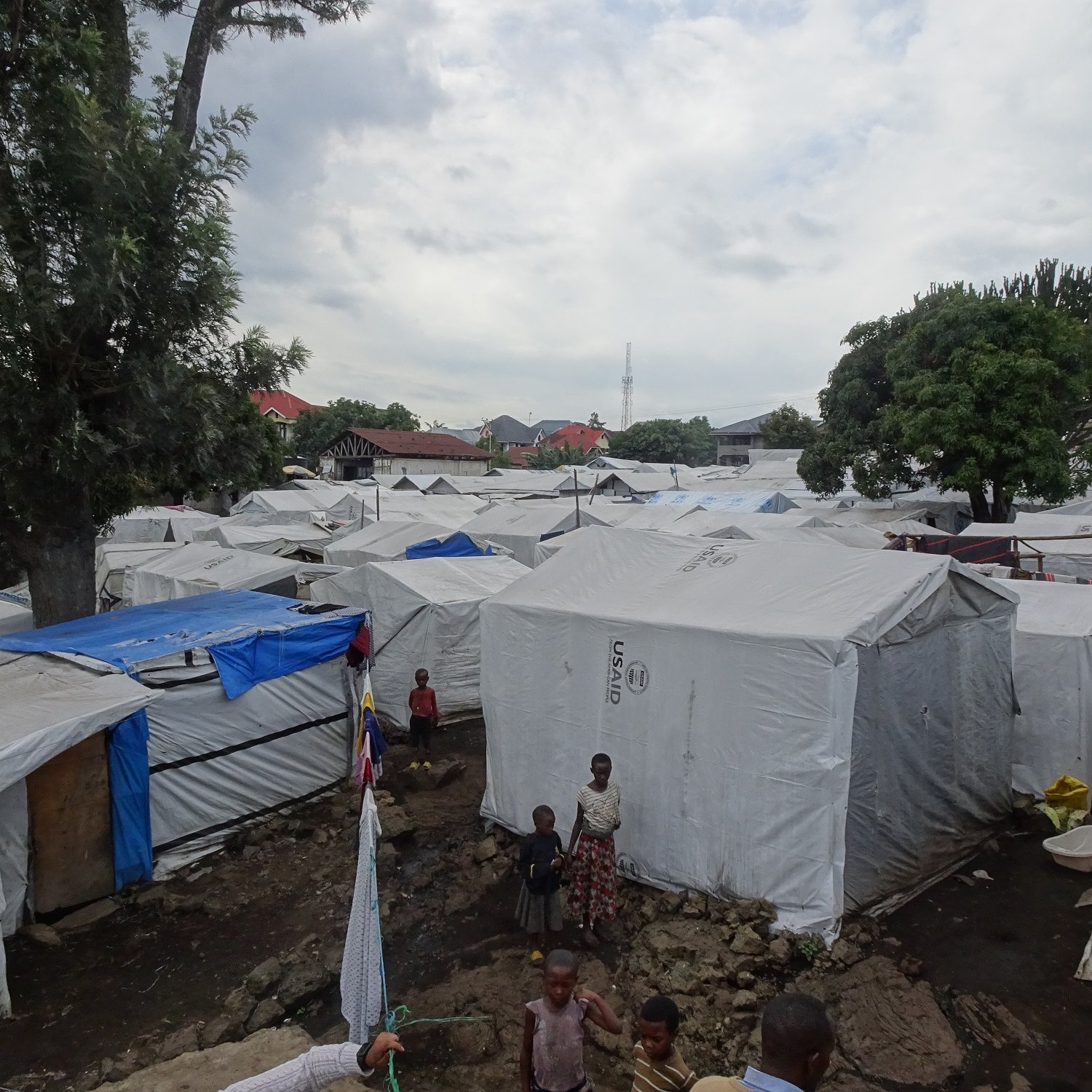 Cette image montre un paysage d'un camp de réfugiés, avec de nombreuses tentes blanches disposées de manière un peu désordonnée. Les tentes sont recouvertes d'une toile claire qui les protège des intempéries. On aperçoit quelques enfants jouant au milieu des tentes. Au loin, des arbres abondants ajoutent une touche de verdure, et on peut distinguer quelques structures plus solides. Le ciel est nuageux, suggérant qu'il pourrait pleuvoir, ce qui souligne la vulnérabilité du cadre. L'ambiance générale semble à la fois animée et calme, tandis que le sol est irrégulier, avec des pierres et des zones boueuses.