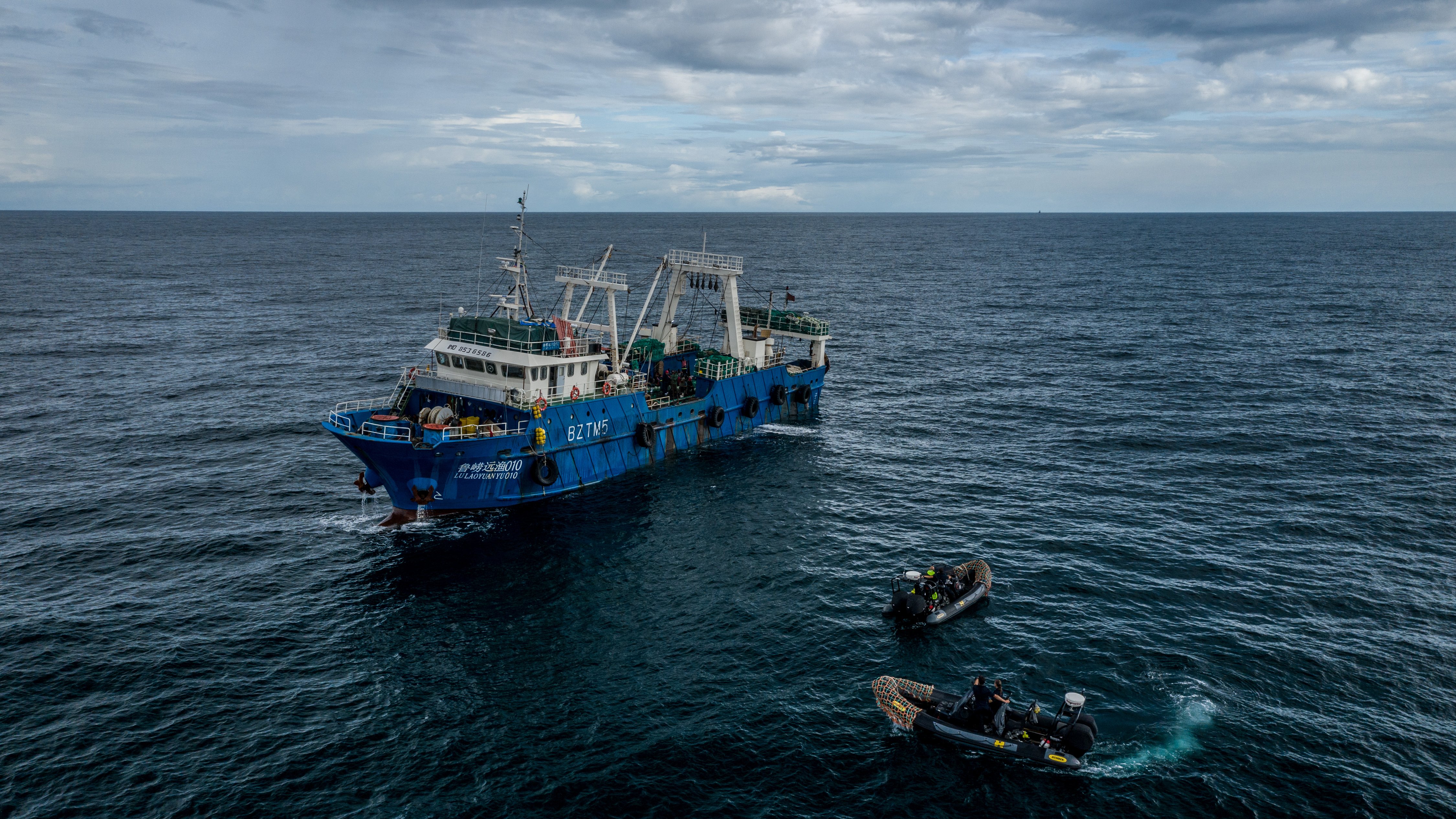 L'image montre un grand navire de pêche de couleur bleue, flottant au milieu de l'océan. Le bateau est en mouvement sur des eaux agitées, sous un ciel nuageux qui laisse présager une légère brise. À côté du navire principal, on peut voir deux petites embarcations, probablement des canots, qui sont en train de s'approcher. Les membres d'équipage, habillés en tenues de plongée, semblent occupés à des activités liées à la pêche. L'ambiance générale dégage une impression de travail maritime, avec le son des vagues et un léger souffle du vent.