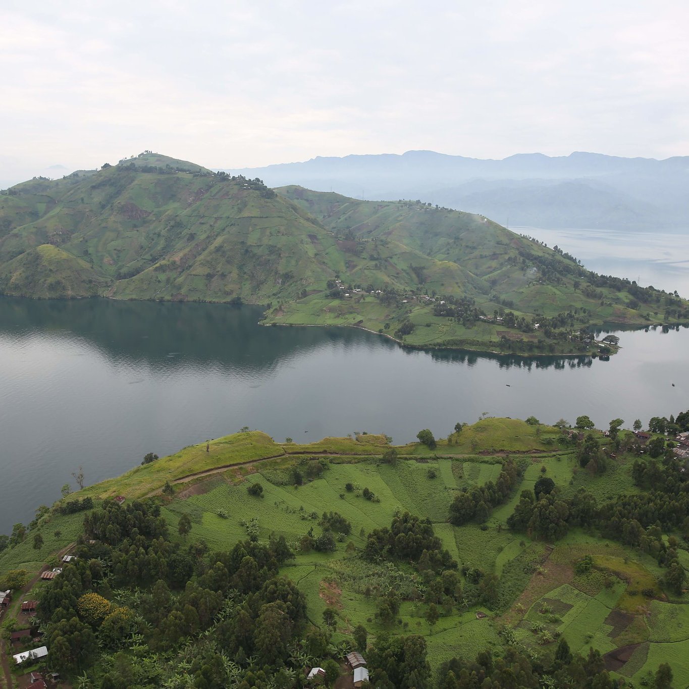 Imaginez un vaste paysage naturel serein, où les collines verdoyantes et les rives d'un lac s'étendent à perte de vue. Au premier plan, des champs verdoyants semblent onduler sur le sol, parsemés de petites habitations rustiques. En arrière-plan, une immense colline se dresse majestueusement, recouverte de nuances de vert qui varient avec la lumière. Le lac, calme et réfléctif, capture le ciel gris clair au-dessus. À l'horizon, d'autres collines se dessinent, créant une impression de profondeur et de tranquillité dans ce cadre apaisant. L'ensemble dégage une atmosphère de paix et de beauté naturelle, accentuée par le murmure léger de l'eau.