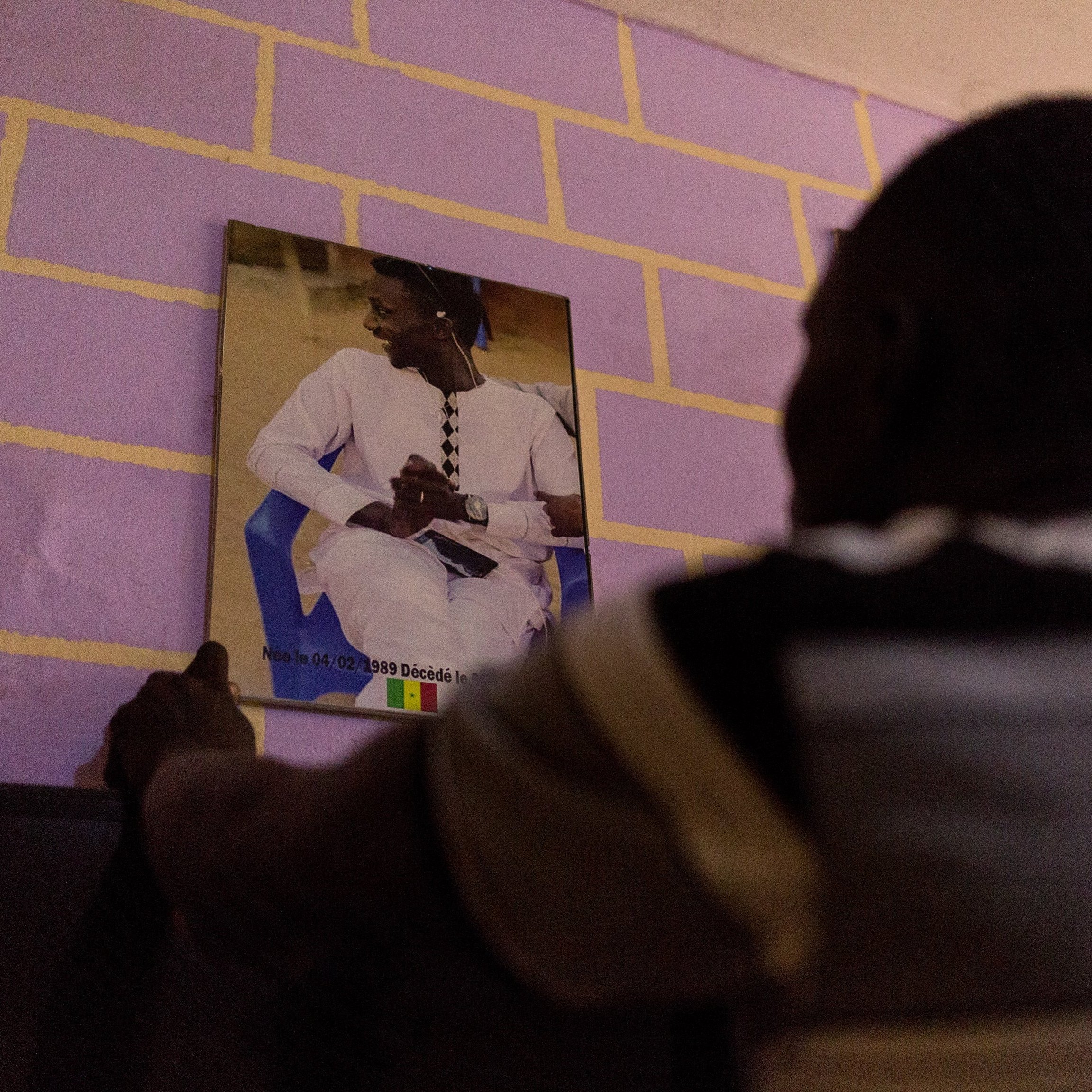 Dans cette image, un homme est en train de fixer un portrait au mur. Il se tient de dos, face à un mur peint en violet aux briques jaunes. Le portrait montre une personne assise, vêtue d'une tenue traditionnelle, probablement sénégalaise, avec un fond flou. L'homme qui fixe le portrait semble concentré sur sa tâche, ajoutant une touche personnelle à l'espace où il se trouve.