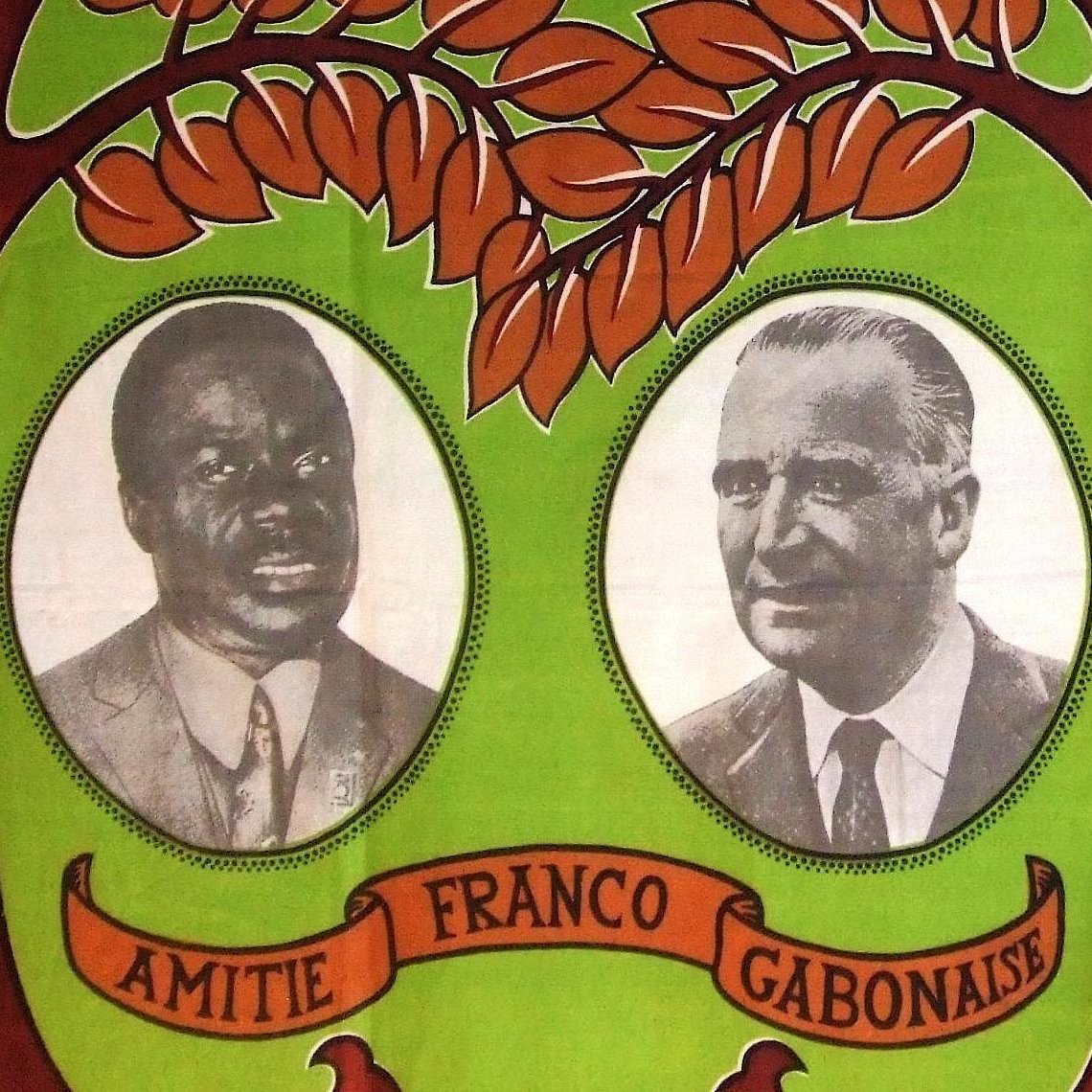 L'image présente un fond aux couleurs vives, dominé par des teintes vertes et marron. Au centre, deux portraits en noir et blanc sont encadrés. À gauche se trouve le portrait d'un homme à la peau noire, coiffé d'une chemise claire et affichant un regard sérieux. À droite, un homme à la peau claire, vêtu d'une chemise sombre, a une expression face à la caméra qui évoque la confiance. Entre les deux portraits, le mot "AMITIE" est inscrit en lettres majuscules, suivi de "FRANCO GABONAISE", soulignant un lien symbolique. Le motif de feuilles stylisées et d'ornements naturels en arrière-plan évoque un environnement tropical et chaleureux, symbolisant l'unité et la collaboration entre les cultures.
