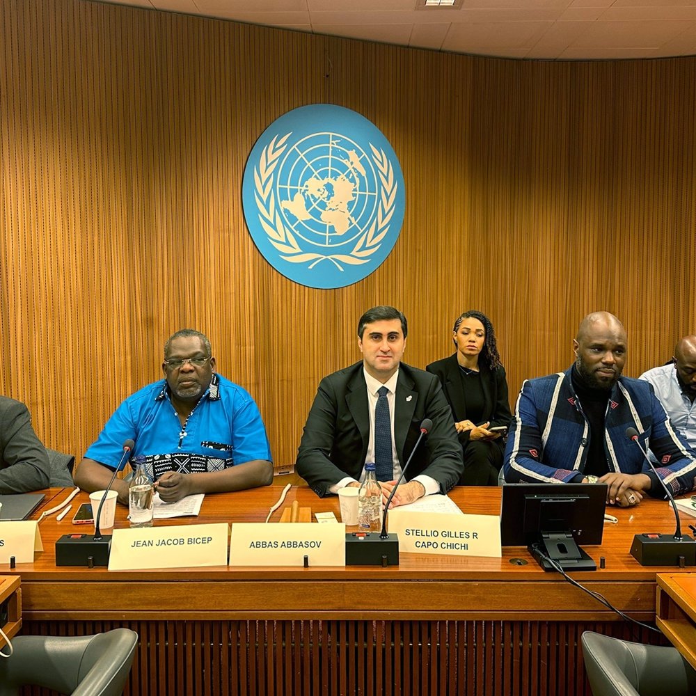 L'image montre une scène à une table de conférence, probablement lors d'une réunion aux Nations Unies. Au centre, un homme en costume noir et cravate blanche, avec un air sérieux, semble être le porte-parole ou le modérateur. À sa gauche, un homme portant une chemise bleue et des lunettes, avec un air attentif. À sa droite, un homme d'apparence plus décontractée avec un pull et une barbe, qui semble également participer à la discussion. En arrière-plan, plusieurs personnes sont assises, écoutant attentivement. Le décor est sobre, avec des murs en bois et le logo des Nations Unies en arrière-plan.