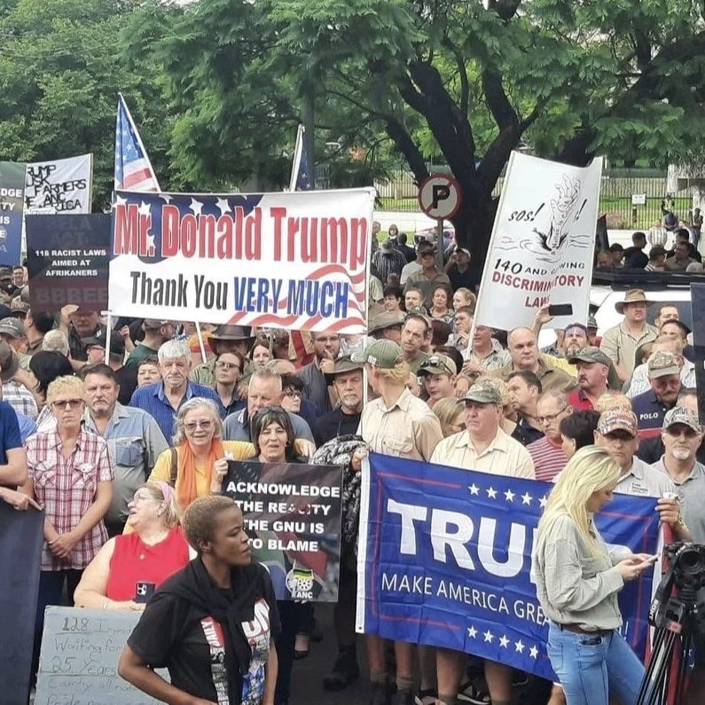 L'image montre une grande foule rassemblée lors d'une manifestation. Les participants tiennent divers panneaux et banderoles. Certains panneaux expriment leur soutien à Donald Trump avec des messages comme "Thank you VERY MUCH" et "MAKE AMERICA GREAT AGAIN". D'autres affichent des revendications ou des slogans liés à des préoccupations politiques. Les manifestants présentent une variété d'âges et de styles vestimentaires, certains portant des chapeaux emblématiques. L'atmosphère semble animée, avec des expressions de passion sur leurs visages. En arrière-plan, des arbres et des panneaux de signalisation sont visibles, créant un cadre urbain pour cette manifestation.