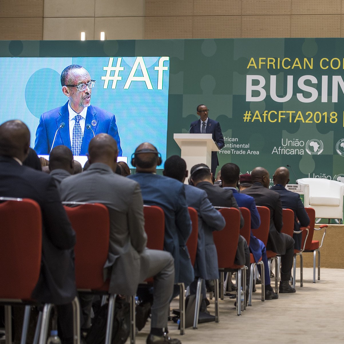 L'image présente un forum d'affaires qui se tient dans le cadre de la Zone de libre-échange continentale africaine. Au premier plan, on voit un homme, vraisemblablement un orateur, debout au micro, s'adressant au public. Il est entouré d'une scène bien agencée avec plusieurs chaises blanches et des logos des organisations impliquées dans l'événement. Le public, de dos, est composé d'hommes en costume, attentifs à la présentation. L'ambiance semble formelle et axée sur les discussions économiques. En arrière-plan, des banderoles mentionnent le forum et sa date, soulignant l'importance de l'événement pour le commerce en Afrique.