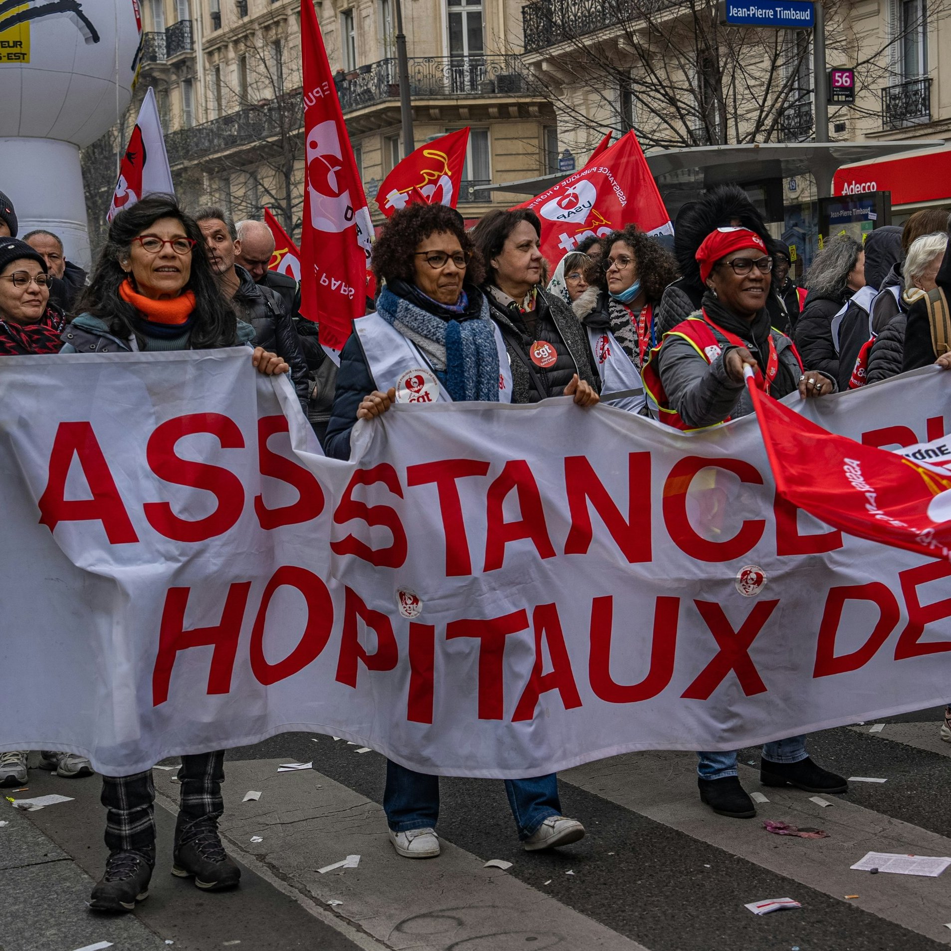 L'image montre une manifestation dans une rue animée. Au premier plan, un groupe de personnes tient une grande banderole blanche sur laquelle est écrit "ASSISTANCE HÔPITAUX DE…" en lettres rouges. Les manifestants arborent des sourires et semblent déterminés. On aperçoit également plusieurs drapeaux rouges, qui sont souvent associés à des syndicats. L'atmosphère est dynamique, avec une multitude de personnes autour, représentant la solidarité et l'engagement. Les bâtiments en arrière-plan suggèrent une ambiance urbaine, typique des grandes villes.