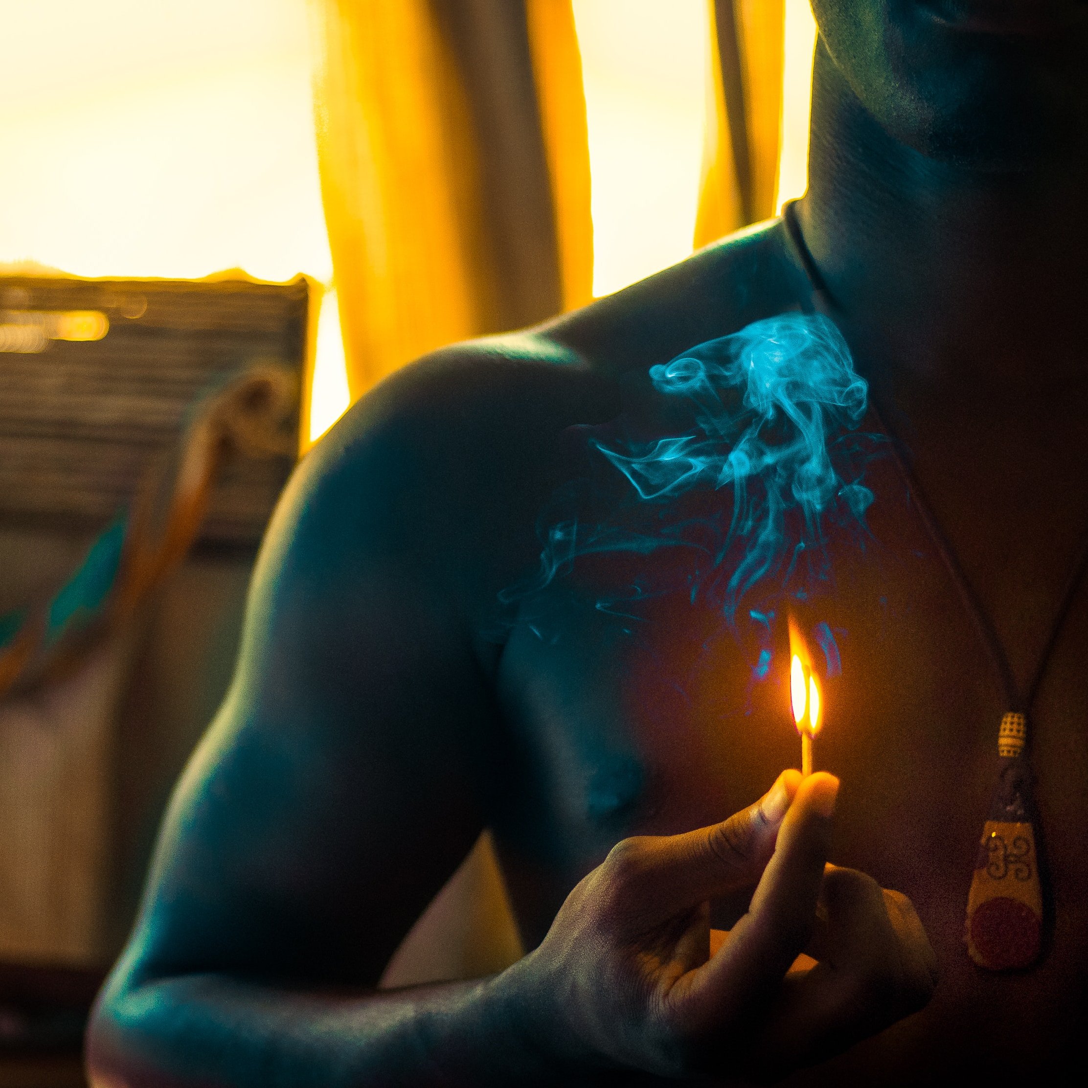 Dans cette image, nous voyons un homme torse nu tenant une petite flamme d'une allumette qui fume légèrement. La lumière douce qui l'entoure crée une ambiance chaleureuse. À l'arrière-plan, des éléments flous suggèrent un environnement intérieur, peut-être une pièce éclairée par le soleil couchant, ce qui ajoute une touche de chaleur à l'atmosphère. La fumée s'élève lentement, créant un contrasté avec la lueur de la flamme.