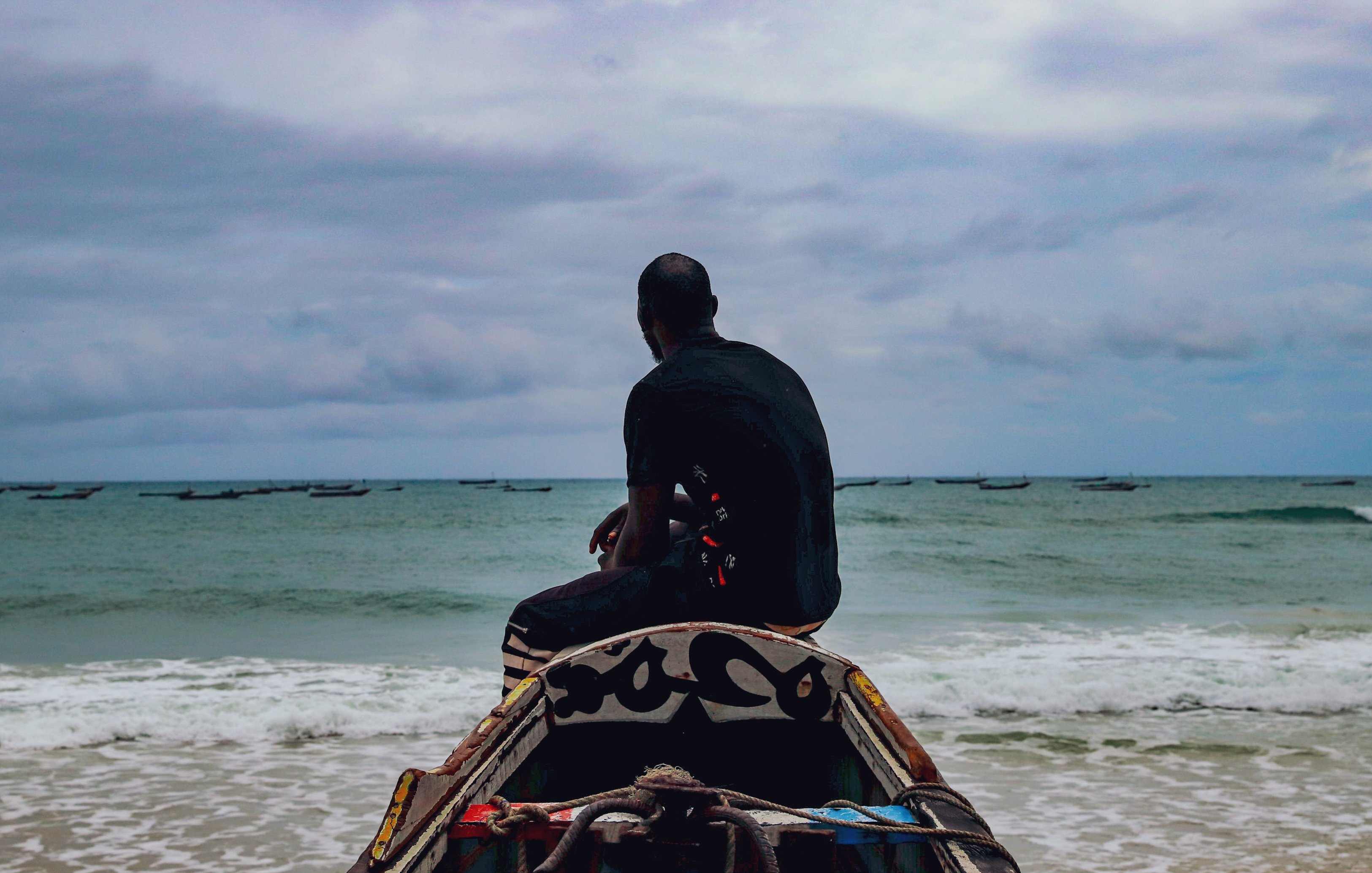L'image montre un homme assis sur une barque, tourné vers la mer. Il regarde l'horizon, où l'eau rencontre le ciel nuageux. La mer est calme avec quelques petites vagues, et on aperçoit des bateaux au loin. L'ambiance est sereine, soulignée par le ciel gris et la lumière douce. L'homme porte un vêtement sombre et semble pensif, contemplant la vastitude de l'océan devant lui.