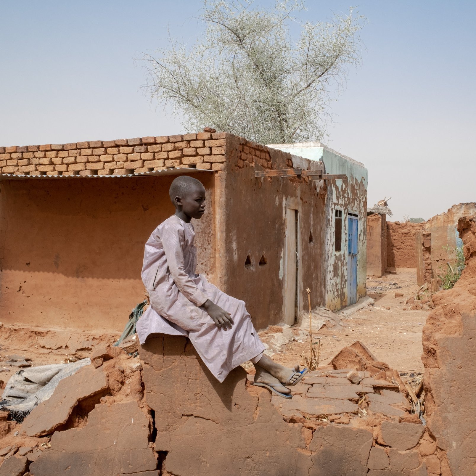 L'image montre un jeune garçon assis sur un mur de terre, au milieu d'un paysage rural aride. Il porte une longue tunique claire et semble pensif. En arrière-plan, on voit des maisons en terre battue, avec des murs qui sont fissurés ou partiellement détruits. L'environnement est sec, avec quelques arbres isolés. Le ciel est dégagé, d'un bleu clair. L'atmosphère générale transmet une sensation de solitude et de résilience dans un milieu rural difficile.
