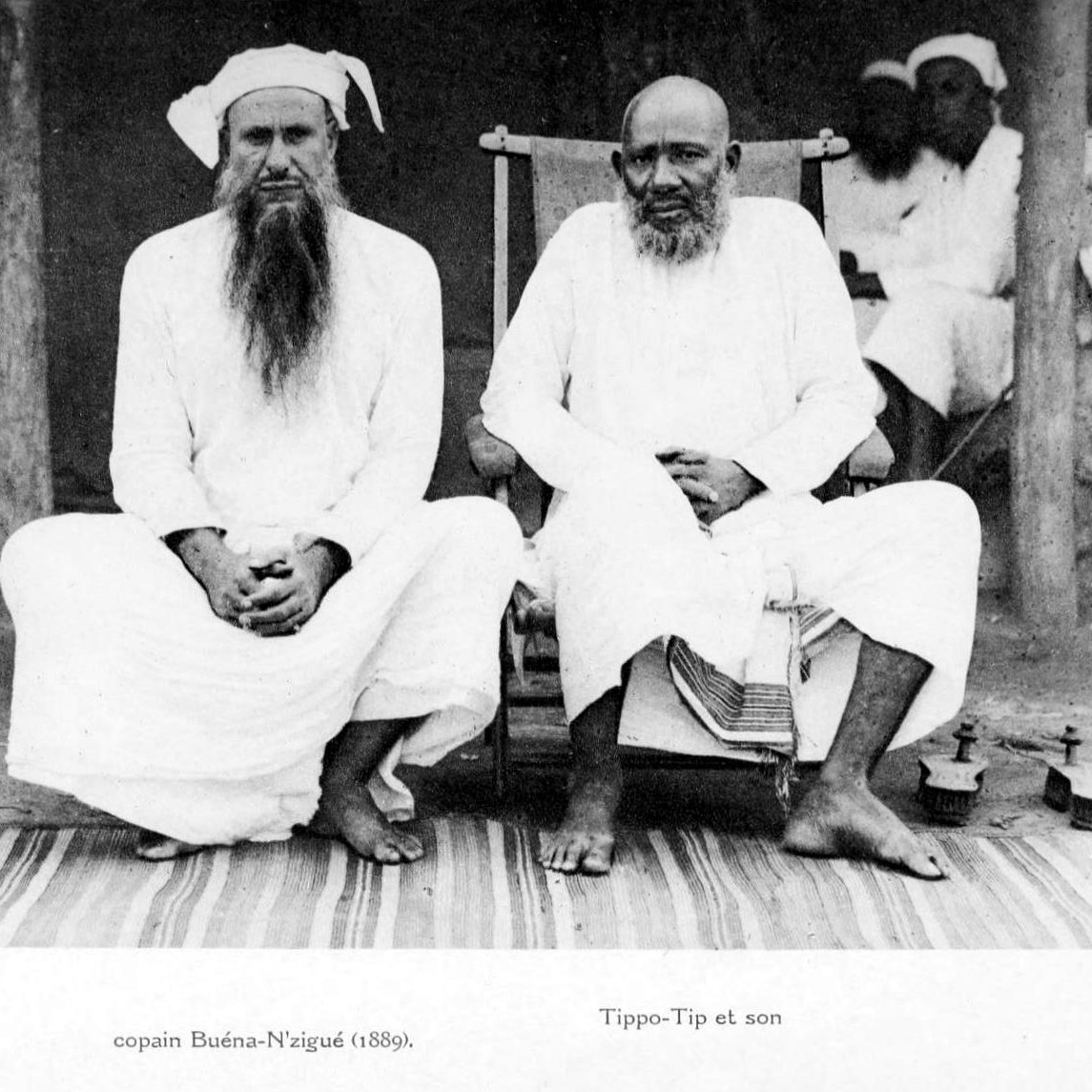 L'image montre deux hommes assis côte à côte sur un tapis au sol. L'homme de gauche, portant une longue barbe blanche et un turban, est habillé d'une tunique blanche. À sa droite se trouve un autre homme, également en tunique blanche, qui semble plus âgé, avec une barbe grise. Ils affichent tous deux une expression calme et sereine. En arrière-plan, on peut distinguer des silhouettes d'autres personnes floues et un environnement rural. Ces détails donnent une impression de tranquillité et de tradition.