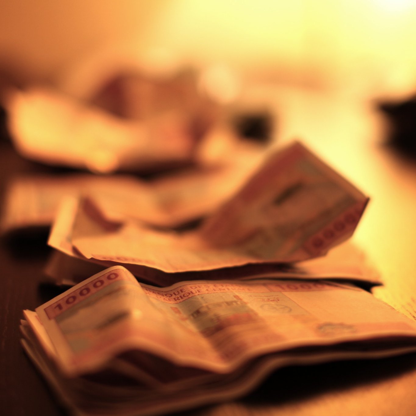 L'image montre un plan rapproché de billets de banque étalés sur une surface sombre, probablement une table en bois. Les billets sont flous, suggérant une légère profondeur de champ, ce qui attire l'attention sur la texture et les détails des billets. L'éclairage est doux et chaud, avec une lumière diffuse provenant d'une source hors du champ de vision, créant une ambiance intime et feutrée. Cette lumière fait ressortir les motifs et les couleurs des billets, ajoutant une dimension visuelle à l'ensemble.