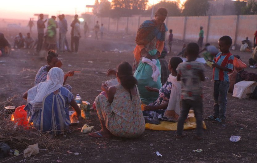 L'image montre une scène de vie en plein air, probablement à la tombée de la nuit. Au premier plan, plusieurs femmes et enfants sont assis sur le sol, entourés de quelques objets personnels et de restes de nourriture. Les femmes, habillées de vêtements colorés, semblent préparer un repas autour d'un feu. En arrière-plan, la lumière du crépuscule illumine légèrement la scène, et de nombreuses personnes peuvent être aperçues se déplaçant dans un environnement poussiéreux. L'atmosphère dégage à la fois une certaine convivialité et une sensation de précarité.