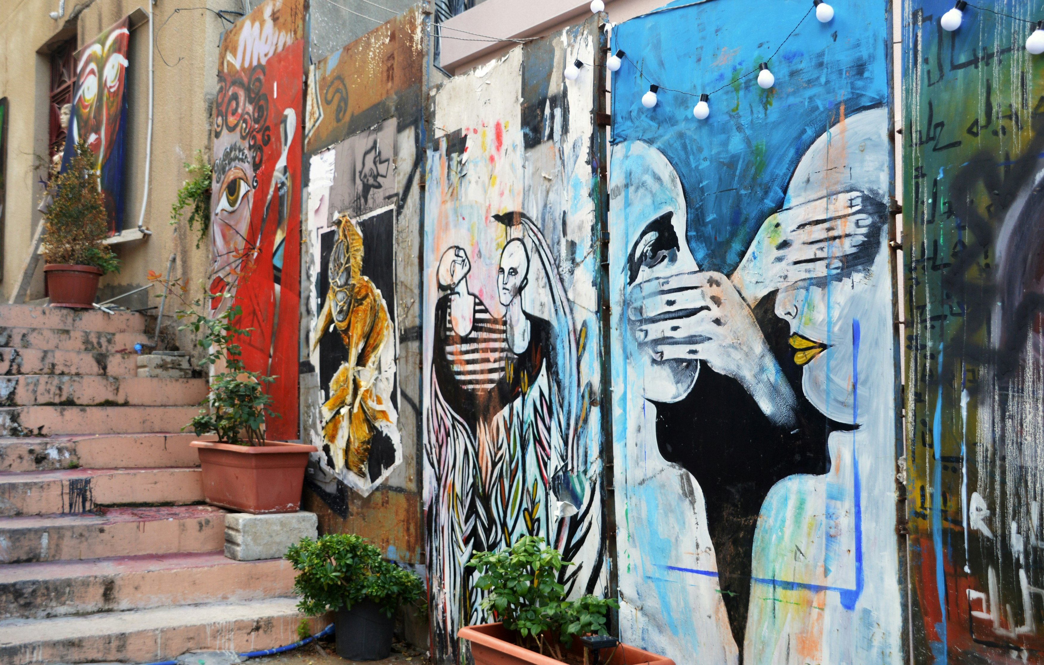 L'image présente un mur de graffitis colorés qui ornent une ruelle. On peut distinguer plusieurs œuvres d'art, chacune ayant son propre style. À gauche, une figure regardante avec des détails réalistes, suivie d'illustrations plus abstraites et expressives au centre et à droite, représentant des visages et des mains en mouvement. Les couleurs dominantes incluent des nuances vives de bleu, d'orange et de noir. Des pots de fleurs, avec des plantes vertes, ajoutent une touche de nature à l'environnement urbain. Au-dessus des œuvres, des guirlandes lumineuses créent une ambiance chaleureuse. Les escaliers en pierre accompagnent le mur, suggérant une connexion avec l'espace environnant. L'ensemble dégage une atmosphère artistique et dynamique, reflet d'un lieu vivant et créatif.