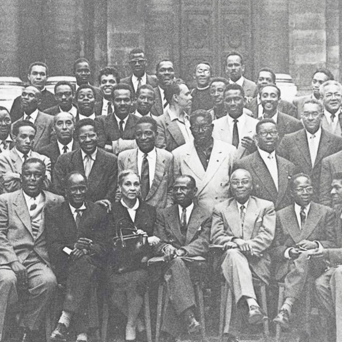 L'image présente un groupe de plusieurs hommes, probablement des leaders et des intellectuels, rassemblés pour une photographie de groupe. Ils sont vêtus de costumes formels, avec une variété de poses : certains sont assis, d'autres debout. L'ambiance semble chaleureuse et conviviale. Au centre, une femme se distingue, assise avec un sourire. En arrière-plan, on aperçoit un bâtiment historique, suggérant peut-être un événement ou une rencontre importante. L'image est en noir et blanc, ce qui lui donne un aspect intemporel.